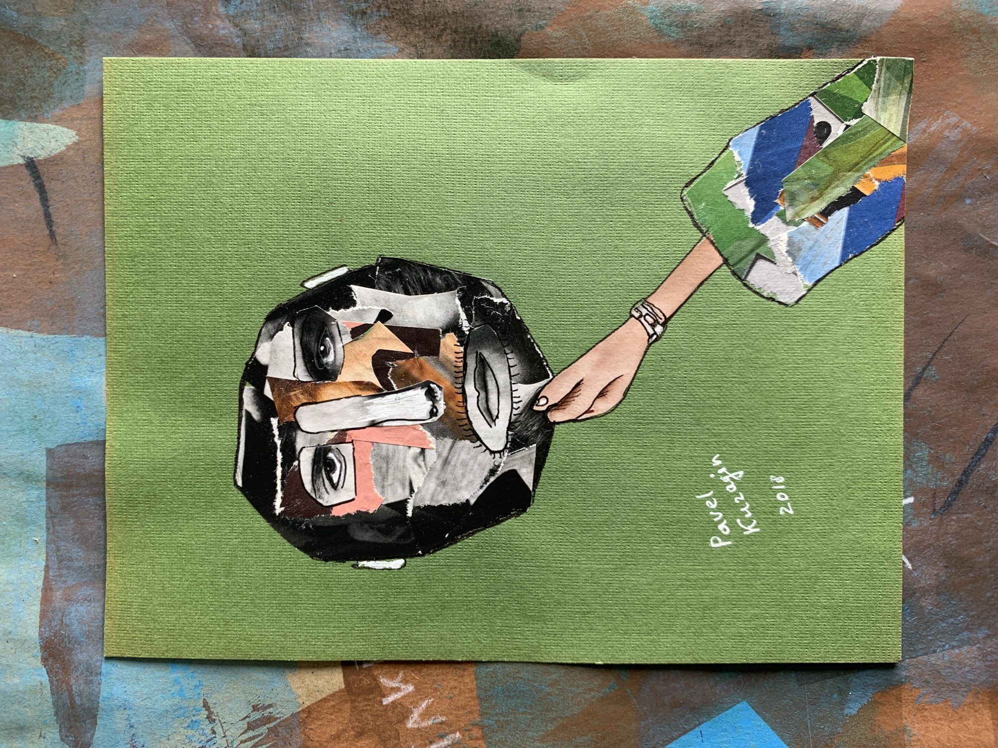This is a collage artwork on a green textured paper. The central subject is a stylized, abstract face created from ripped pieces of paper. The face has a round, somewhat bulbous shape and incorporates various materials, including black and white photographic elements, flesh-toned paper for skin, and what appears to be parts of a tire for texture. It features two eyes, one made from a circular cutout with black lines, and another from a collection of smaller paper fragments. A light pink piece of paper is visible near the nose area, and a silver-toned object, possibly a wristwatch, is depicted on the wrist of a hand.
A disembodied hand, rendered in a light peach skin tone with black outlines, extends from the right side of the image. The hand is adorned with a silver bracelet. This hand is holding or interacting with a collage element in the shape of a shield or flag, composed of fragmented pieces of colored paper, predominantly blues, greens, and some yellow and orange. This element is attached to the right side of the artwork.
Below the hand and to the right, handwritten text in white is visible. It reads "Pavel Kuzagin" and "2018." The background of the artwork is a solid, textured green paper. The overall impression is a surreal and abstract portrait, possibly exploring themes of identity or perception through the juxtaposition of diverse materials. The activity depicted is ambiguous, but the extended hand suggests interaction or presentation of the collage element. There are no explicit cues regarding time of day, weather, or a specific location. The artwork conveys a sense of artistic experimentation and creative expression.