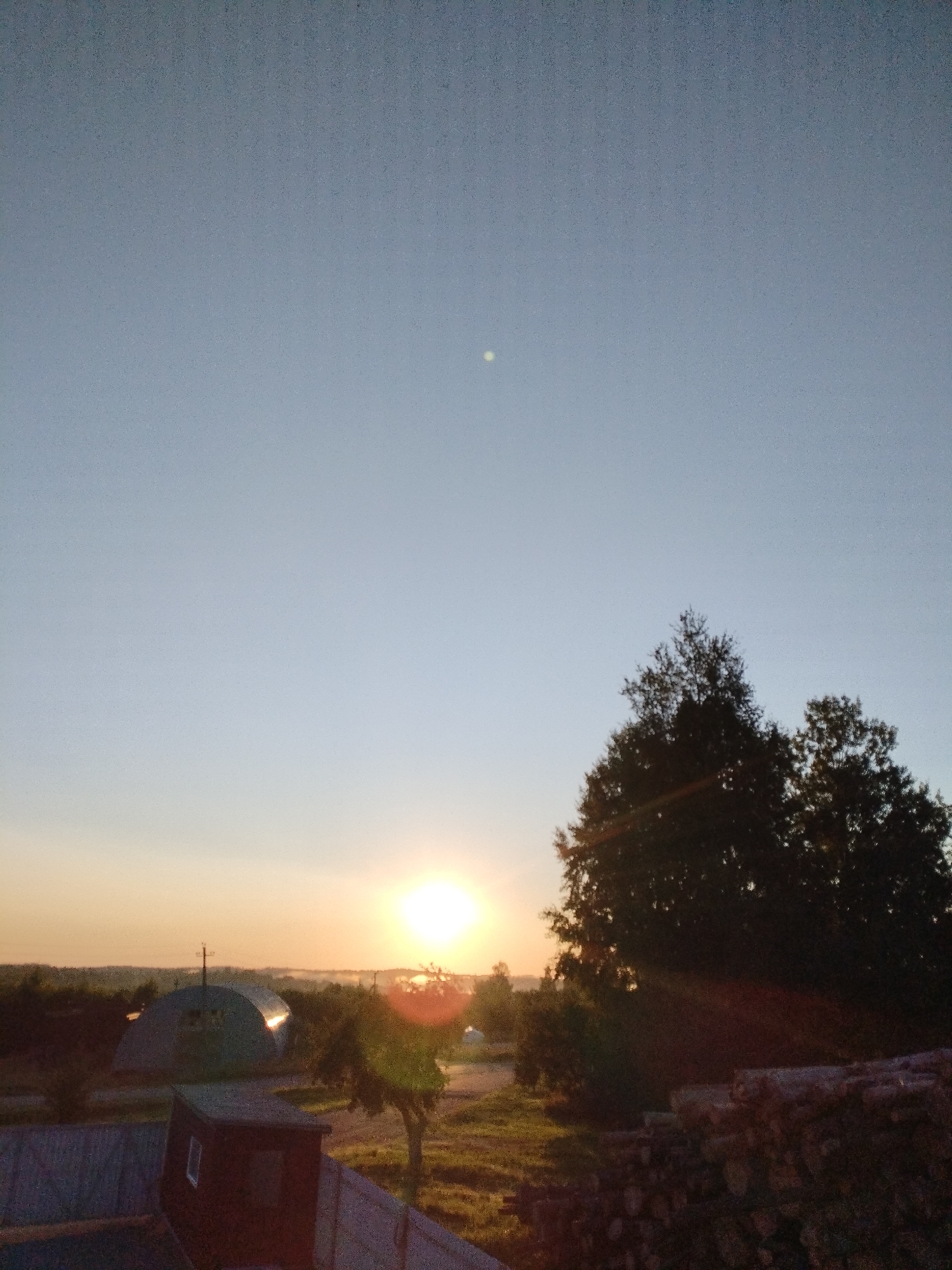 A rural landscape is depicted at dusk, with the sun setting in the background. A large, arched structure, possibly a barn or hangar, is visible in the middle ground. In the foreground, a wooden fence and a stack of firewood are present. The sky is clear and pale blue, with a faint light in the upper portion. The image is likely taken in the Вароніцкі сельскі Савет, Belarus, given the rural setting. There are no people visible in the image.