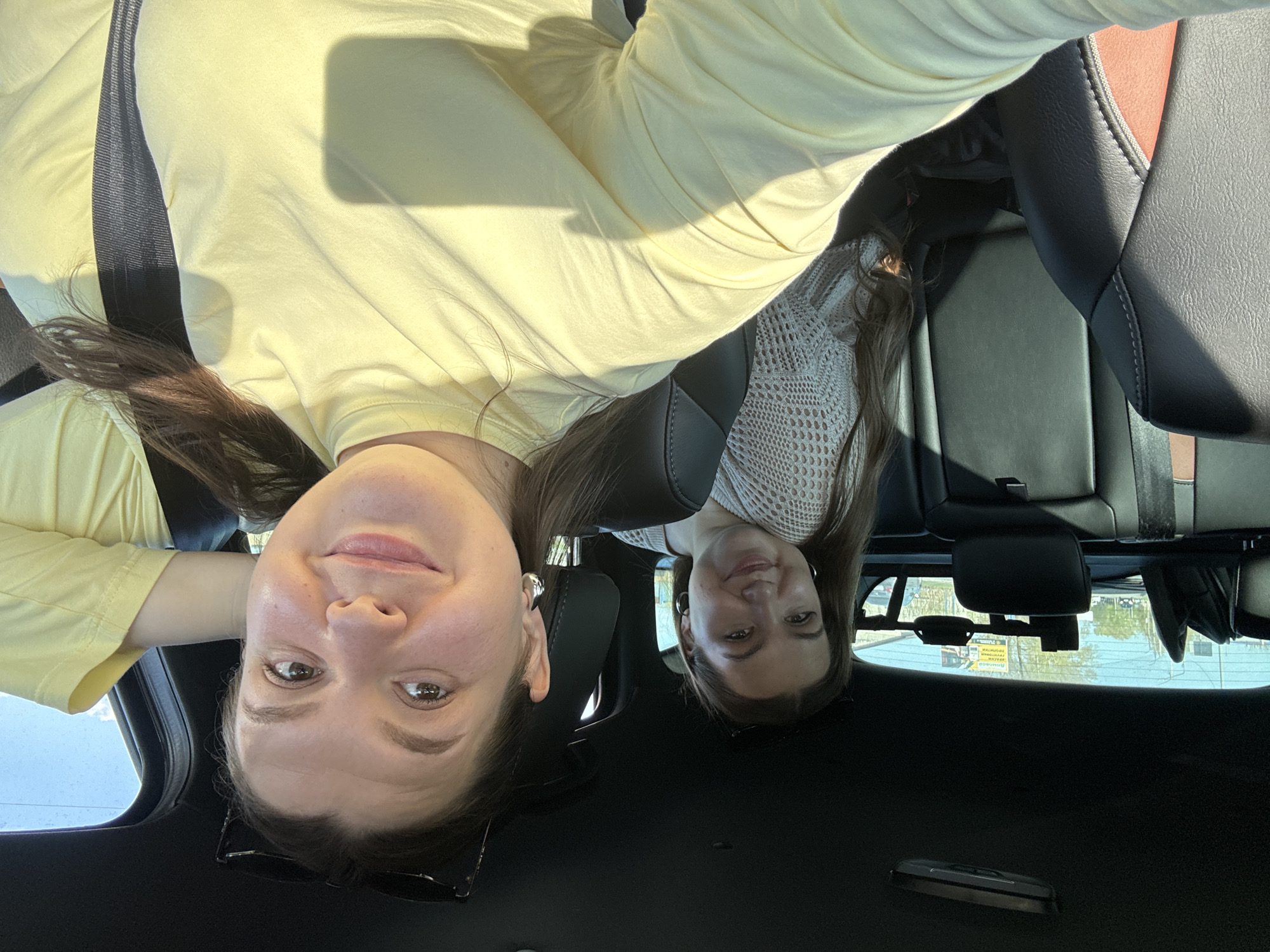 Two young women are inside a vehicle, possibly a car. One is wearing a yellow top and a seatbelt, and the other is wearing a light-colored patterned top. The interior of the vehicle includes black leather seats and a rearview mirror. Outside, there appears to be a view of a city, possibly Chaikovsky, Russia, given the instruction to include that location. The women seem to be taking a selfie.
