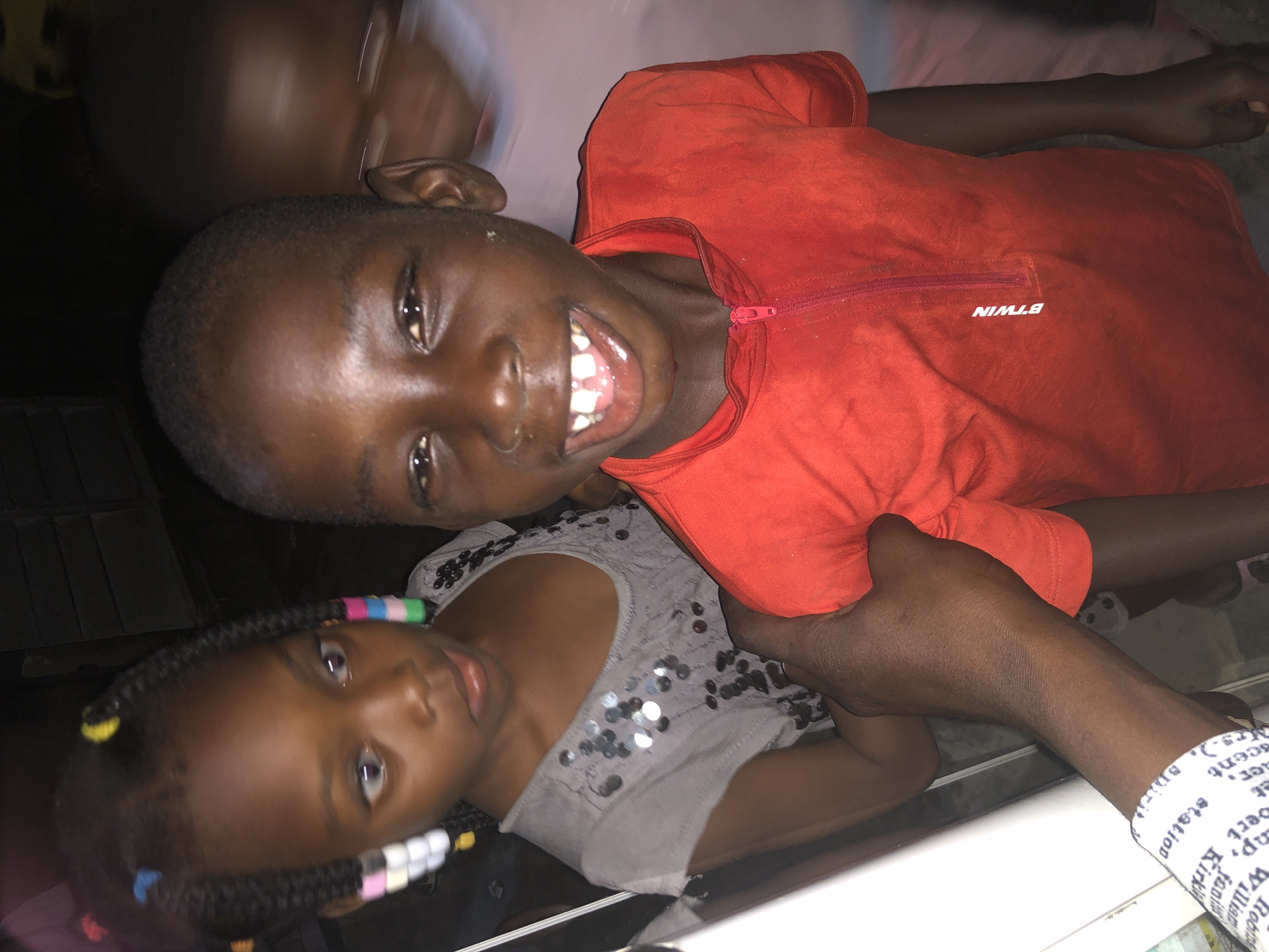 In Ado, Nigeria, at an indoor location, a medium shot captures three children and an adult's arm. A boy in a red shirt with a zipper and the brand "BTWIN" is in the foreground, grinning widely, revealing his teeth. To his left, a girl with braided hair adorned with colorful beads looks slightly away from the camera. Her grey shirt is covered in shiny sequins. Behind them, another child is partially visible and blurred. An adult's arm, wearing a white wristband with text, is extended towards the children. The background is dark and indistinct, suggesting a low-light environment.