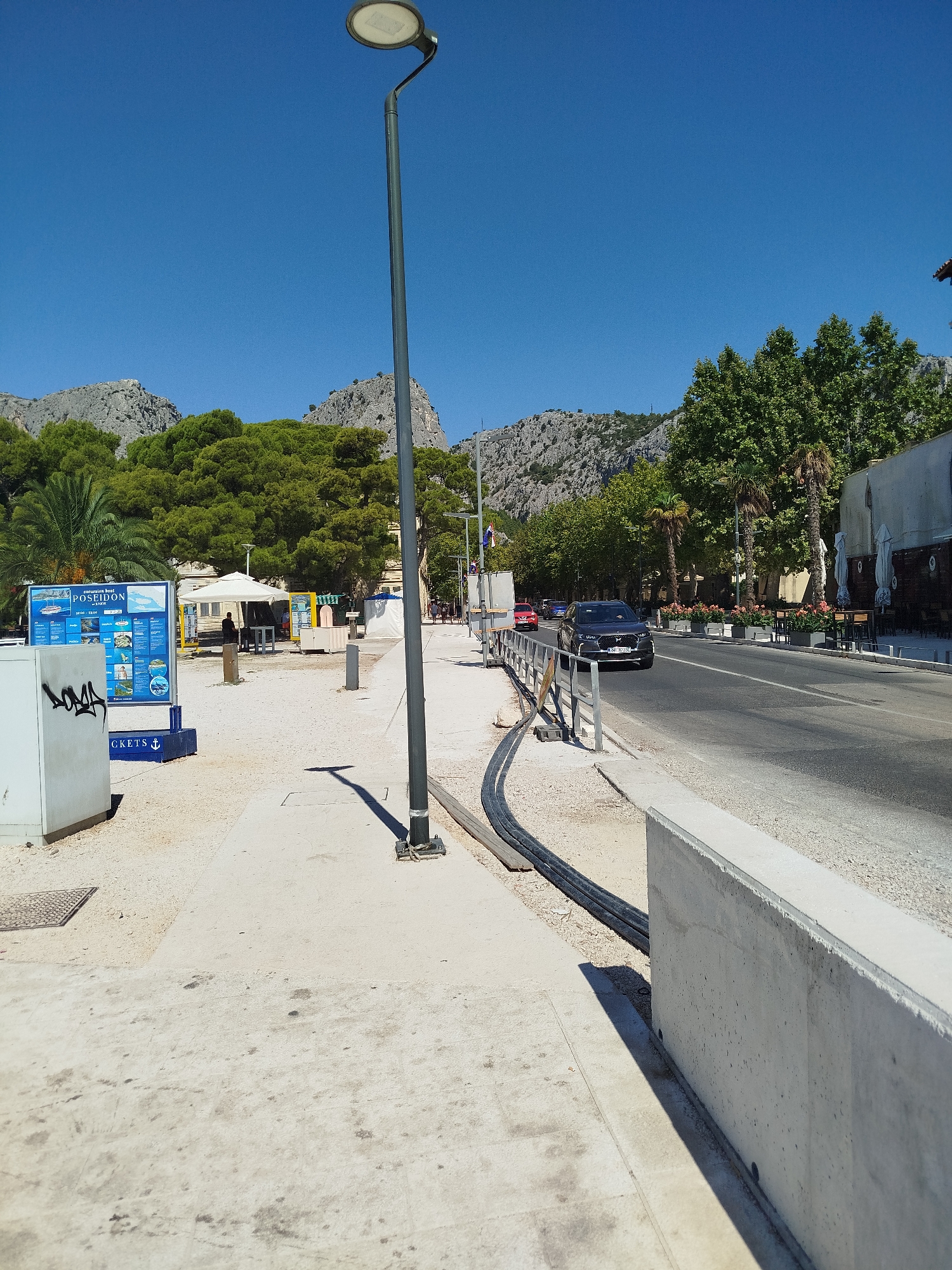 The image depicts a street scene in Omiš, Croatia, with a clear blue sky overhead. In the foreground, a concrete sidewalk runs alongside a street, with a tall lamppost positioned prominently. To the left of the sidewalk, a white kiosk displays information, including a sign for "POSEIDON" excursion boats and "TICKETS". A dark-colored SUV is driving on the street, and behind it, a red car is also visible. Trees line the street, and in the background, rocky hills rise under the bright sky. The scene suggests a coastal town environment, possibly a promenade or a busy street near a tourist area.