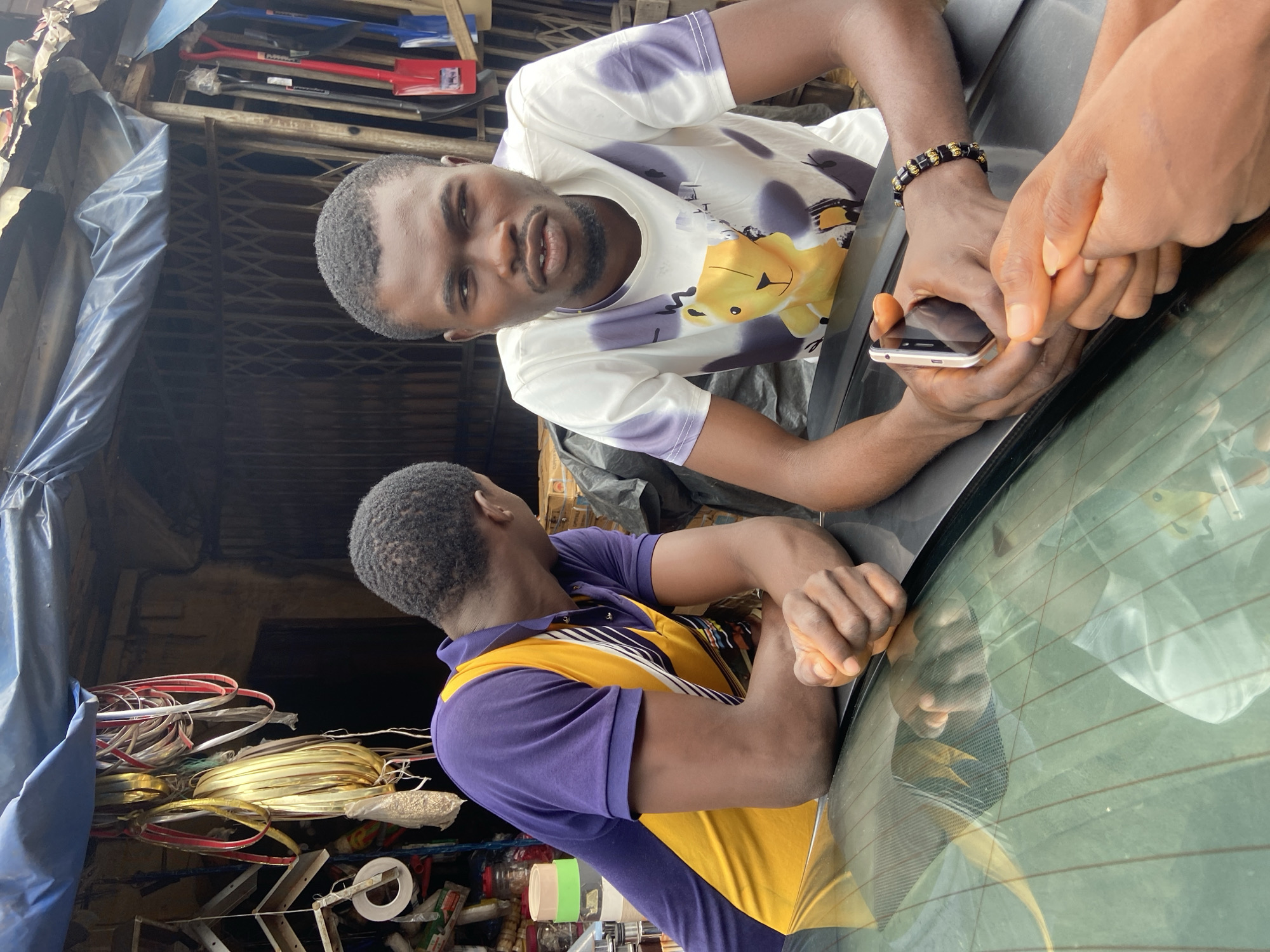 The image shows three individuals in what appears to be an outdoor market setting in Ogidi, Nigeria. The foreground features the rear window of a vehicle, with the reflections of the sky and surrounding environment visible. Two men are prominently featured, leaning over the car.
The man on the left, seen from the back, wears a purple and yellow collared shirt. He is leaning forward, his arms resting on the car. His hair is short and dark.
The man in the center is facing the camera and is the primary subject. He has a short haircut and is wearing a white t-shirt with purple tie-dye patterns and a graphic of a cartoonish yellow animal with a crown. He is looking towards the camera with a neutral expression. His left hand, adorned with a black and gold beaded bracelet, is resting on the car. His right hand is holding a silver smartphone.
A third person's hands are visible on the right side of the frame, also resting on the car, suggesting their presence or interaction with the other two. These hands appear to be holding the man's phone and are reaching towards it.
In the background, a market stall with various items is visible, including rolls of wire (some gold and red, others gold), tools like shovels, and other assorted goods. A blue tarp is also visible, covering some of the market's inventory. The overall scene suggests a busy, possibly informal marketplace environment. The lighting indicates it is daytime. No specific weather conditions are discernible. There is no visible text in the image.
