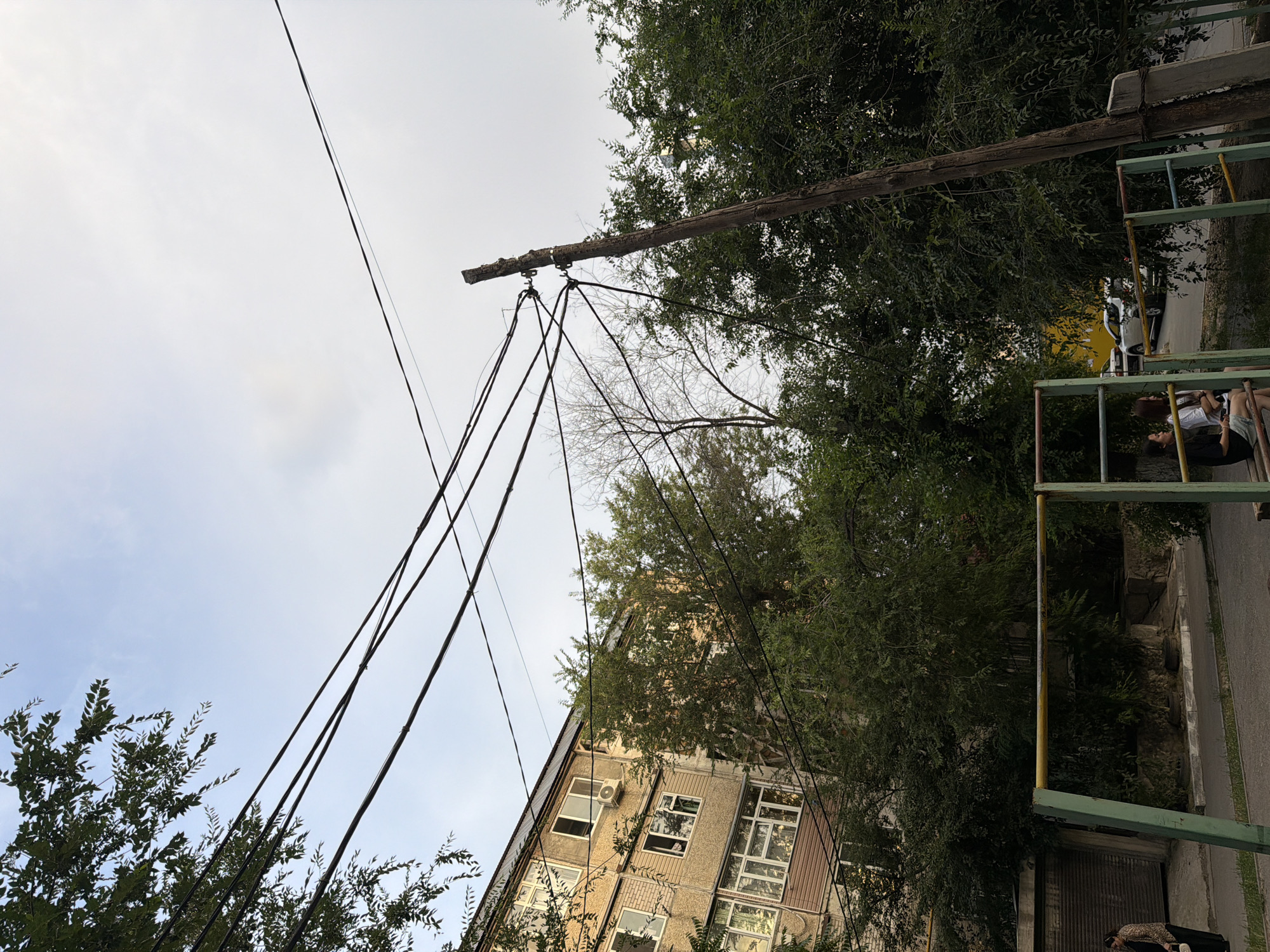 The image depicts an outdoor scene in Shymkent, Kazakhstan, with an apartment building and trees in the background. Power lines are strung across the frame, supported by a wooden utility pole on the right. In the foreground, a playground structure with green and yellow bars is visible. Two young women are seated on the playground equipment, facing each other and appearing to be engaged in conversation. A woman wearing a patterned jacket and dark pants walks past on the left side of the frame. The sky above is overcast.