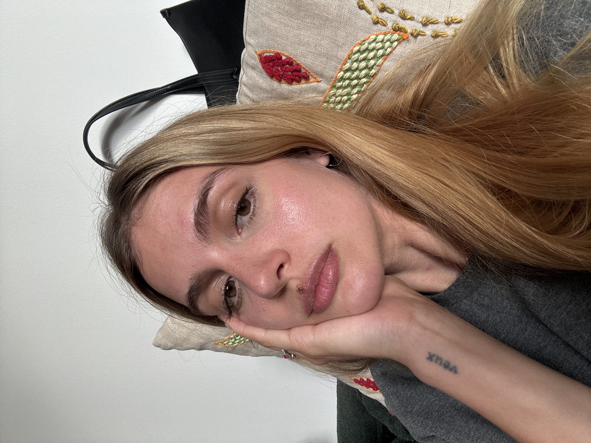 A young woman with long blonde hair is lying down with her head resting on her hand. Her skin appears to be slightly glossy. She is wearing a gray top. On her left wrist is a tattoo that reads "veux." A decorative pillow is positioned behind her head. A black bag is slightly visible in the background. The environment appears to be indoors against a white wall. There are no immediate indicators of a specific city or location.