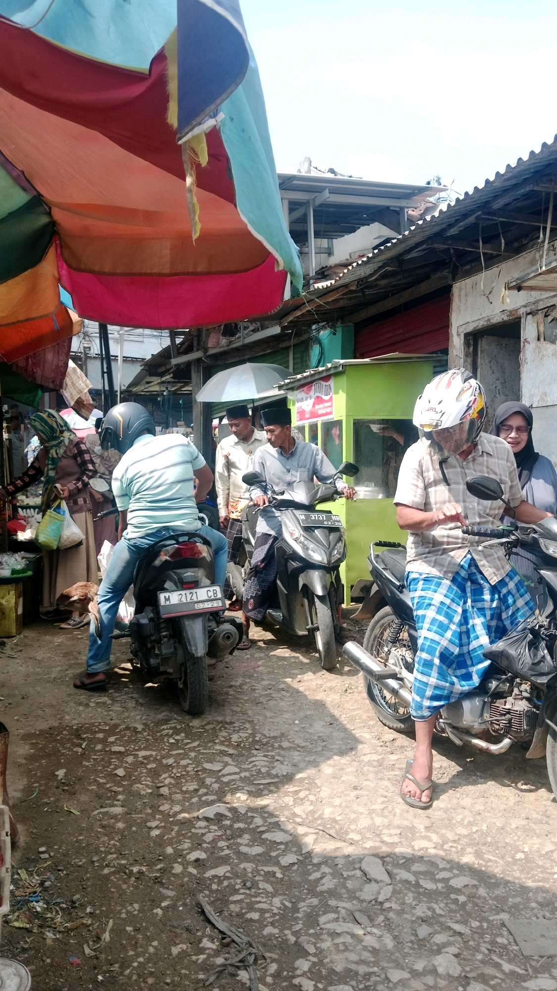The image depicts a street scene in Waru, Indonesia, likely a market or roadside area, under a colorful, patchwork canopy. Several people are present, most on or near motorcycles. In the foreground, a person wearing a white and red helmet and a blue and white checkered sarong sits on a motorcycle, looking to their right. To the left, another person wearing a blue and white striped shirt and blue jeans is seated on a motorcycle with the license plate "M 2121 CB". Behind them, two men stand near motorcycles; one in a light-colored shirt and black cap, and the other in a lighter shirt and black cap. Further back, a woman wearing a hijab and a patterned brown and white top is visible. The environment is somewhat cluttered with debris on the ground, and buildings with corrugated metal roofs are visible in the background. The overall impression is of a busy, everyday street scene.