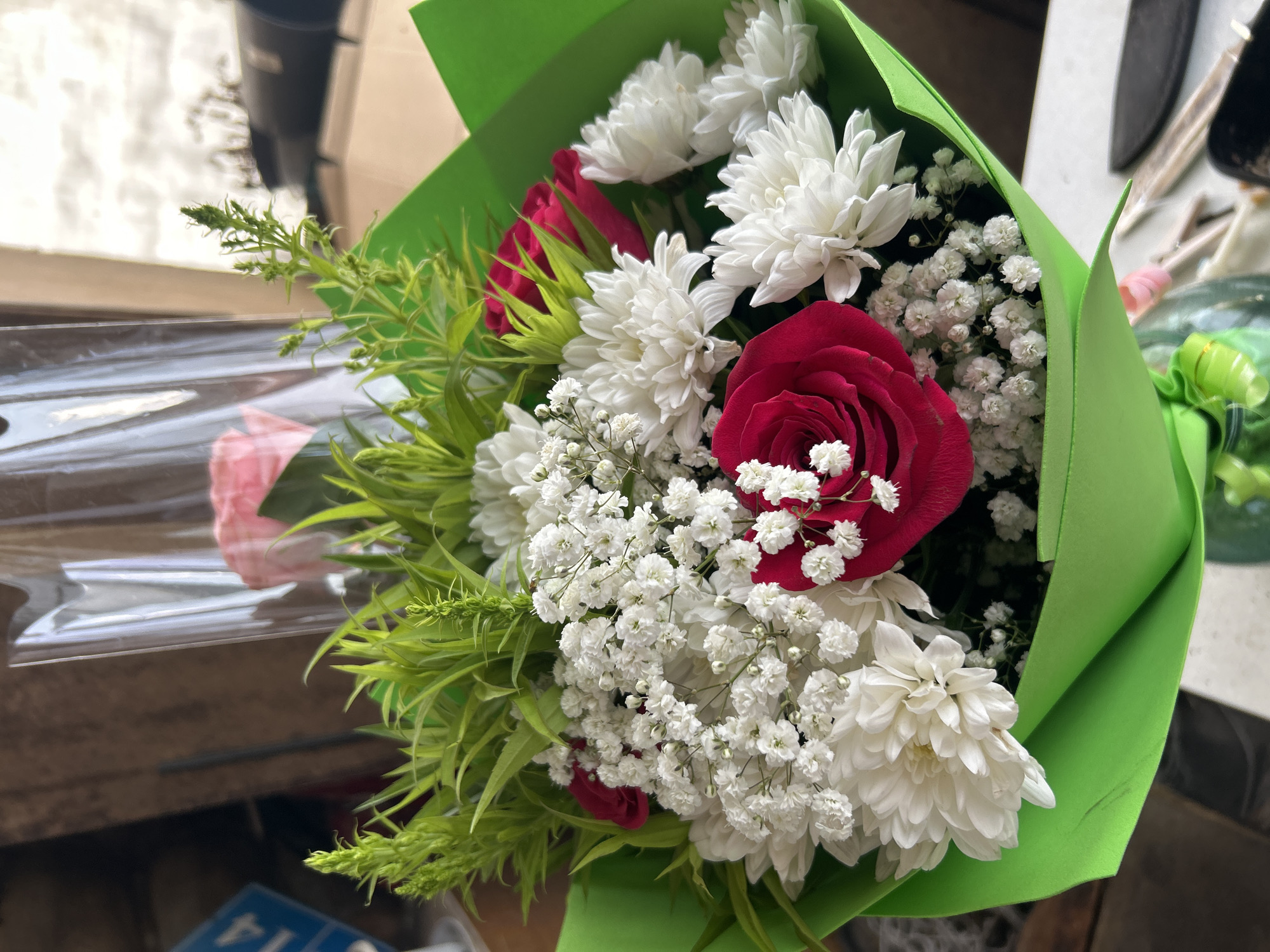 A close-up shot focuses on a vibrant bouquet of flowers, likely red roses, white chrysanthemums, and delicate baby's breath, all wrapped in bright green paper. The bouquet is partially encased in clear plastic. Behind the flowers, the background is blurred, suggesting an indoor setting with wooden elements and a hint of an outdoor scene visible through a window. A blue sign with white numbers, possibly a street address or a price tag, is visible in the upper left corner. The scene is static and appears to be a still life composition. The image does not provide any information about the city of Ignatovo, Russia.