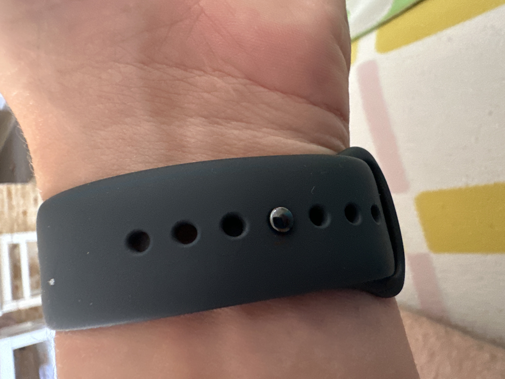 A close-up shot shows a dark grey watchband on a person's wrist. The watchband has a series of circular holes and a silver stud. The skin of the wrist is visible, with some texture and fine hairs. In the background, out of focus, is a pattern of yellow and pink rectangles on a white surface, and a blurred background with white horizontal lines. The image appears to be taken indoors, possibly in a home environment. There is no indication of the city of Budva, Montenegro in the image.