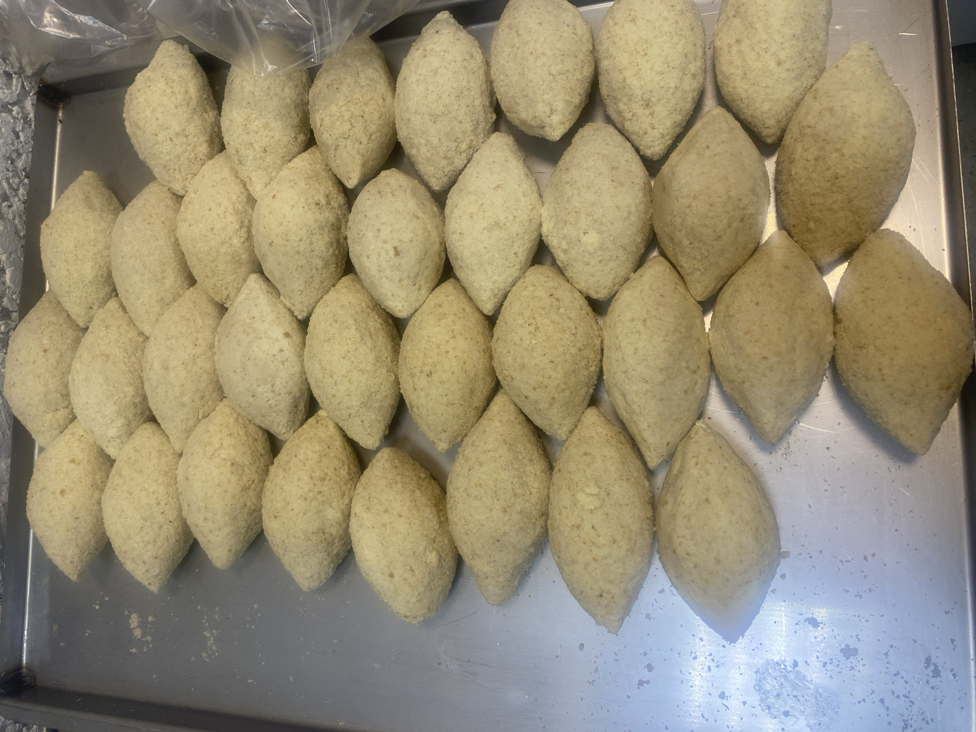 The image shows a tray of approximately 30 kibbeh, arranged in rows and columns. The kibbeh are oval-shaped with pointed ends and are coated in a grainy, beige mixture. They appear to be uncooked. The tray is metal and shows some wear. The background is out of focus. There is no indication of the location or any people present in the image.
