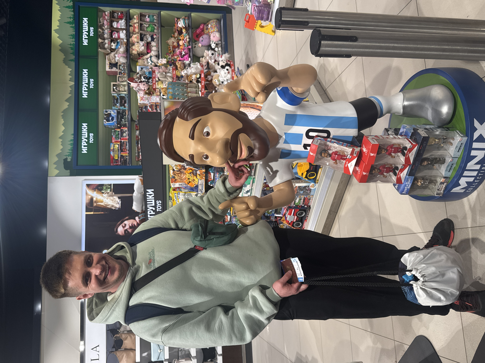 The image shows a man posing next to a life-size cartoon figure of Lionel Messi in a toy store. The man is holding a passport and boarding pass. The toy store shelves display a variety of toys and figurines. The shelves also contain the text "ИГРУШКИ" and "TOYS". The bottom shelves also contain the MINIX collector set. There are no indications this image was taken in Sochi, Russia.