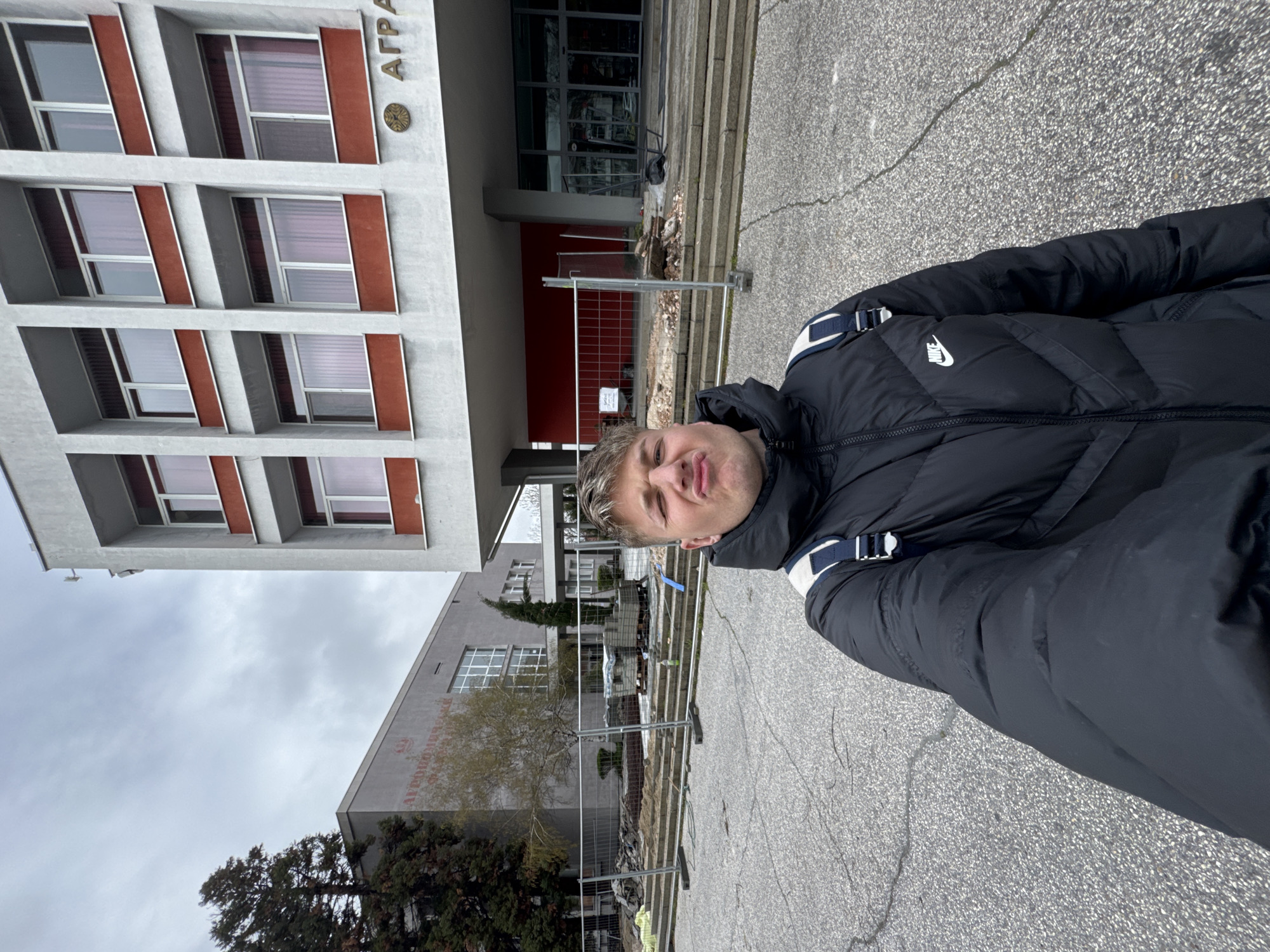 The image shows a man standing outside the Agricultural University of Plovdiv, Bulgaria. He is wearing a black Nike puffer jacket. Behind him, there's a grey concrete building with multiple windows. There are some barriers in front of the building, possibly indicating ongoing construction or repair work. The weather appears overcast.