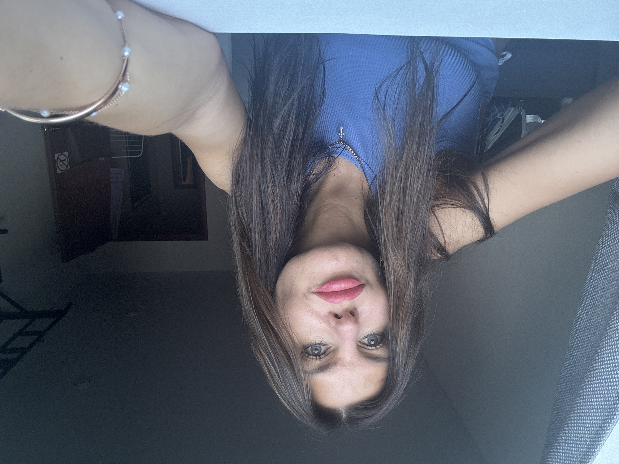 The media file is a selfie taken by a young woman with long, dark brown hair and striking grey-green eyes. She is wearing a blue ribbed tank top and a delicate bracelet adorned with small pearls. A small cross pendant hangs from a thin necklace. Her lips are painted a vibrant pink. The photo is taken indoors, with a dark background that suggests a room with low lighting. To the left, a framed picture and a "no smoking" sign are partially visible. The overall impression is one of casual intimacy, with the woman looking directly at the camera. The selfie appears to have been taken in the afternoon, as indicated by the natural light coming from a window on the right side of the image, which is partially covered by a textured gray curtain. The fact that the image is rotated suggests it was taken by holding the phone or camera at an unusual angle.