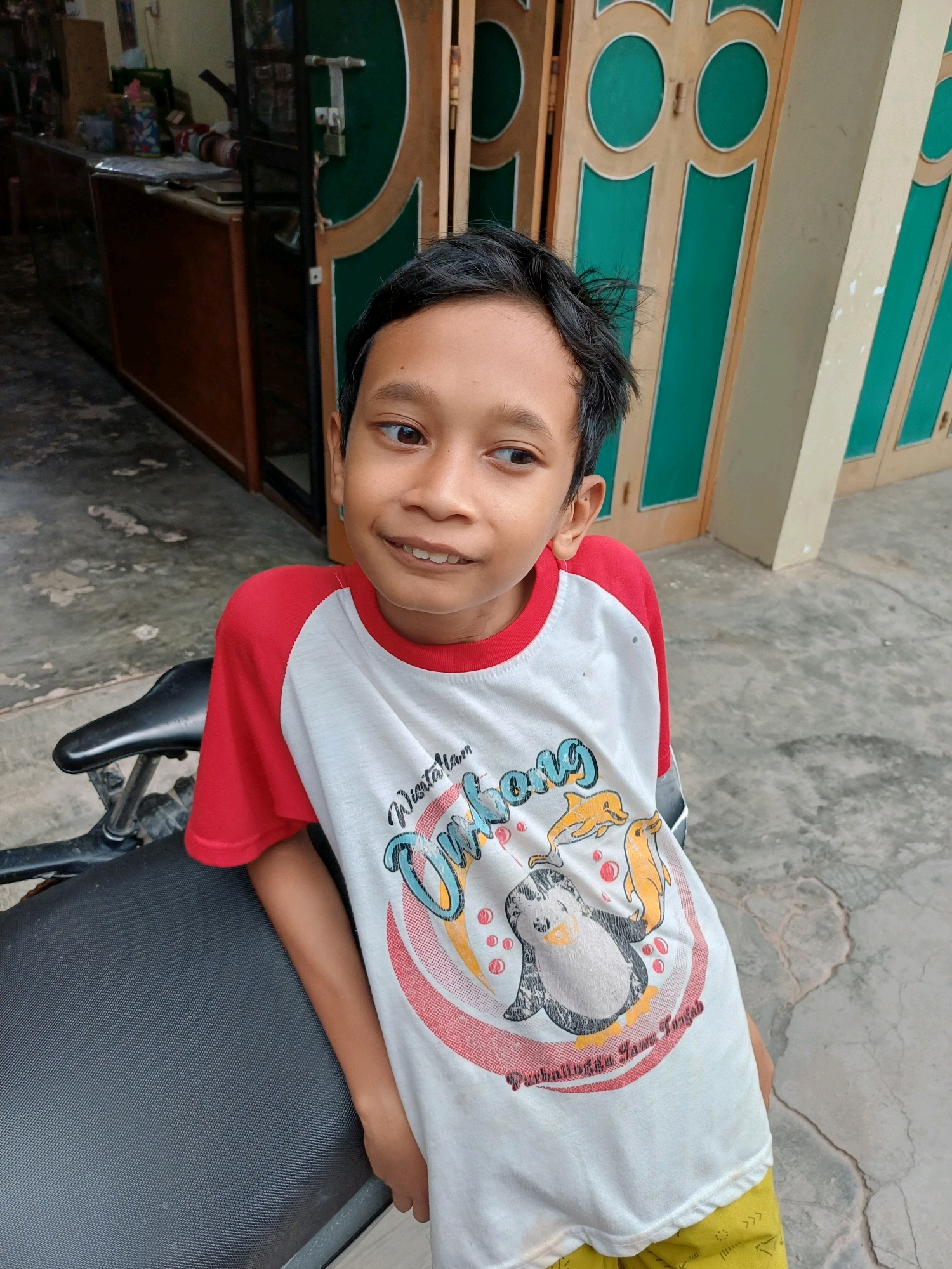 A medium shot, outdoor portrait captures a young boy leaning against the seat of a black motorcycle. The boy is looking to his right and smiling, with his mouth slightly open. He has dark, messy hair and is wearing a white t-shirt with red sleeves. The t-shirt features a graphic print of a penguin and two dolphins inside a circular design, with text above and below. The boy's right arm is resting on the motorcycle seat. The background shows a building with green and wooden doors, and a concrete floor. A wooden counter with various items is visible on the left side of the frame. The setting appears to be a residential or commercial area in Dumai City, Indonesia.