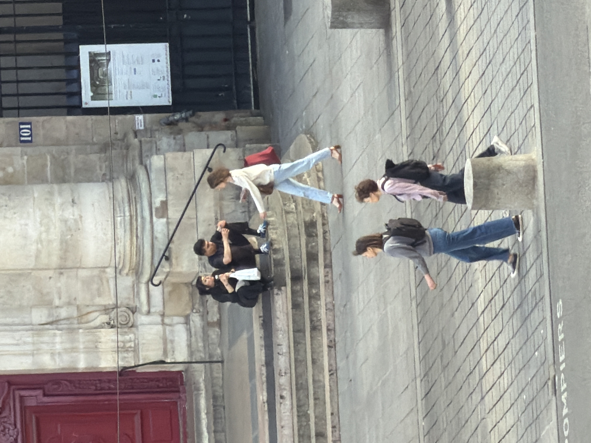 The image captures a bustling street scene in what appears to be a historic urban area, consistent with Paris, France. The scene is bathed in bright daylight, indicating clear weather conditions.
In the foreground, a large, ornate red wooden door or gate is visible on the left. To its right, a wide set of stone steps ascends. Several individuals are present. On the lower steps, two people in dark clothing are seated; one holds a white object, possibly a bottle or bag. Another person in dark attire is positioned slightly above them on the steps. Further up the steps, a person wearing a light-colored long-sleeved top, light blue jeans, and brown shoes is walking.
On the right side of the frame, a paved area with square stone slabs features two more individuals. One person, dressed in a pinkish long-sleeved shirt, dark pants, and a dark backpack, stands near a stone pillar. Another individual, wearing a grey top, blue jeans, and a dark backpack, walks along the paved surface.
In the background, a dark metal gate or railing is visible, behind which a building wall rises. The architectural style, with its stone construction and decorative elements, suggests a historical European context.
Visible text includes a blue plaque with the number "101" affixed to the stone wall above the steps. A white informational sign with text and an image is attached to the metal gate in the upper left. Additionally, the word "POMPIERS" (French for firefighters) is painted in white capital letters on the paved ground in the lower right.