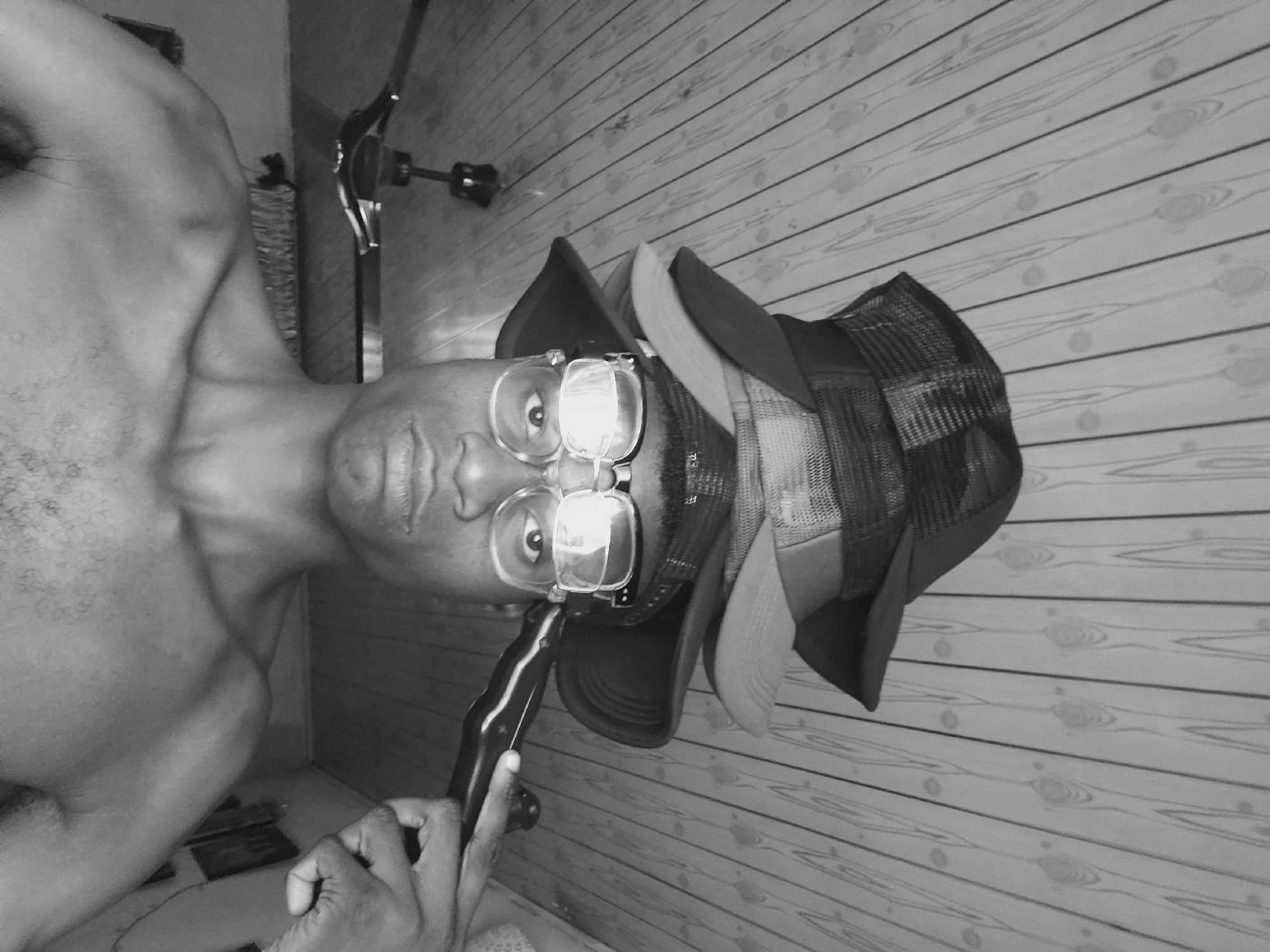 A close-up, black and white image shows a man with a bare chest and multiple hats stacked on his head. He is wearing glasses and holding a black object near his mouth. The background consists of wooden planks running horizontally. The location is unknown, but the image details do not include the city of Narayi, Nigeria.