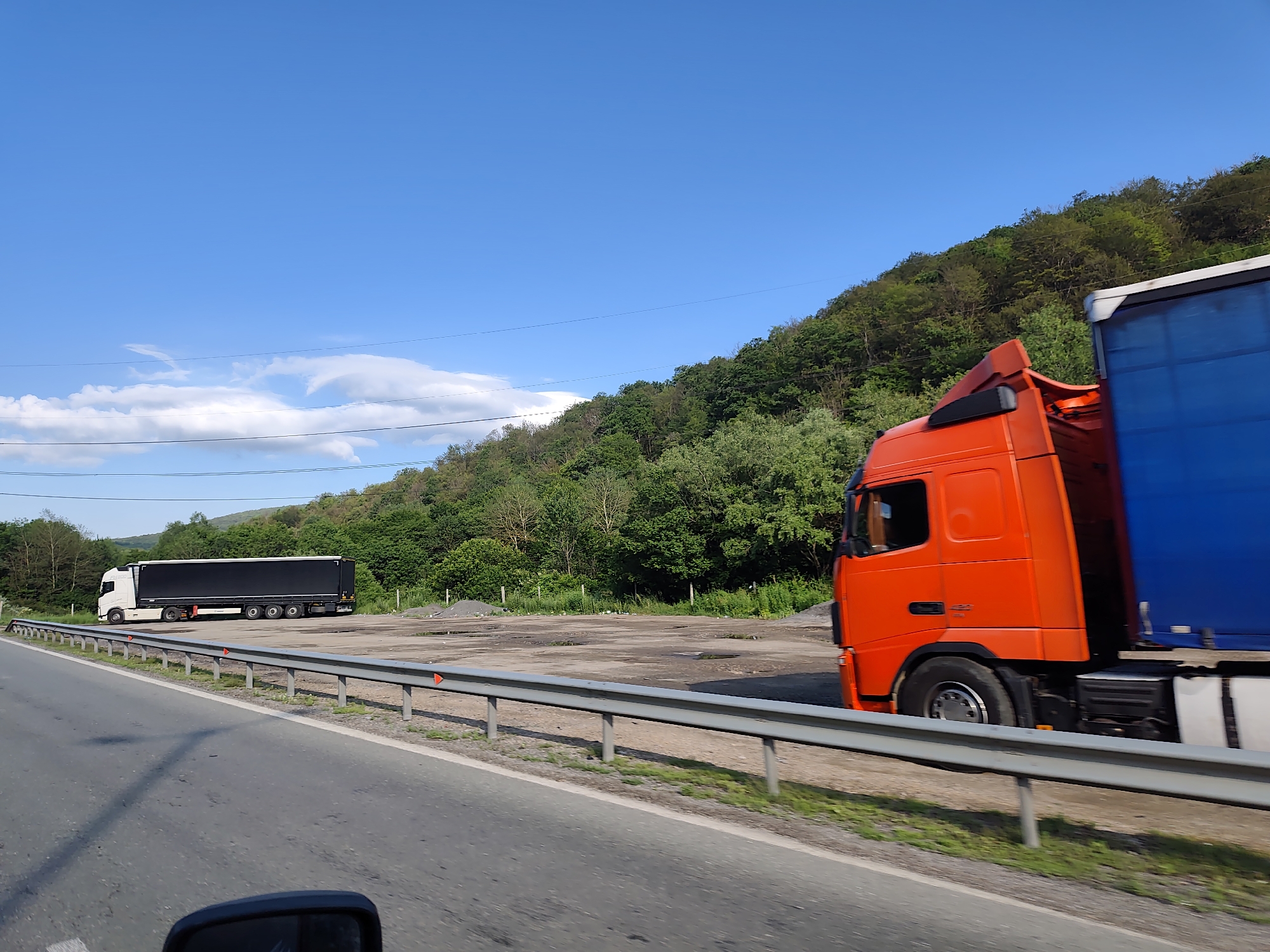 The image captures a roadside scene in Gorno Kamartsi, Bulgaria, on a clear, sunny day. The foreground shows a paved road with a metal guardrail running along its edge. Beyond the guardrail, there is a flat, open area with some patches of gravel and puddles, bordered by a lush green hillside covered in trees. Two large trucks are visible. In the distance on the left, a white truck with a black trailer is parked. Closer to the right side of the image and in motion, an orange truck with a blue trailer is passing by. The sky is a vibrant blue with a few wispy clouds. The overall impression is one of a rural or semi-rural transportation hub.