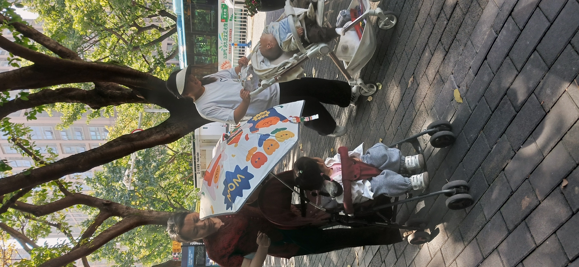 This daytime image captures an outdoor scene in Yuzhong District, China, featuring two women pushing strollers with children on a paved sidewalk. The setting is an urban environment, characterized by large trees with green foliage casting dappled shadows on the dark grey interlocking brick pavement. Tall buildings are visible in the background, hinting at a city landscape.
In the foreground, a woman wearing a red patterned top and dark pants holds a white umbrella adorned with colorful cartoon characters and the word "MOM." She is positioned beside a dark-colored stroller where a toddler, dressed in a pink top, white jacket, and grey sweatpants, sits, appearing to be resting or asleep. Behind her, another woman, wearing a white t-shirt, dark pants, and a white and black visor, pushes a light-colored baby stroller. A baby, clad in a light blue top and dark pants, is visible lying in this stroller.
In the mid-ground, a blue and white bus is seen on a street, featuring Chinese text, including "治气泡九芝堂足光散光" (Treats air bubbles, Jiuzhitang Foot Light Powder) and "汗防复发" (sweat prevention, anti-recurrence), likely an advertisement. Other visible text on signs includes "Public toilet" and "TOURIST." The strong sunlight suggests a clear day, with shadows indicating late morning or early afternoon. The scene conveys a calm and leisurely activity, possibly a walk through a park or pedestrian area.
