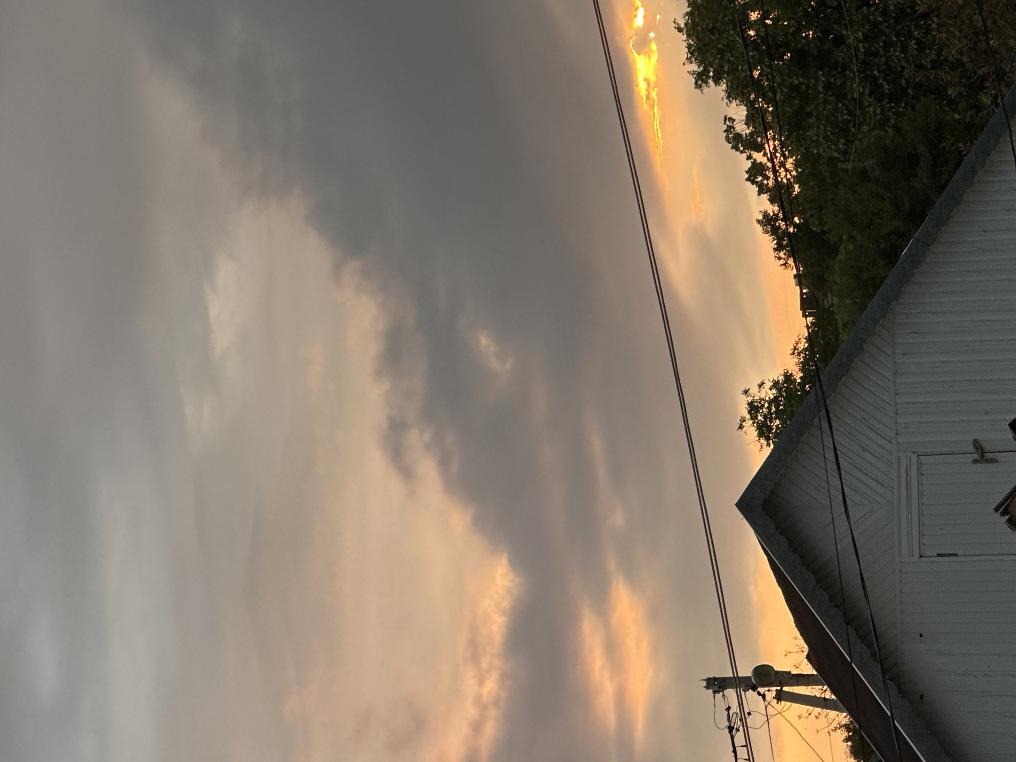 The image captures an outdoor scene at dusk. The sky is filled with a mix of dark gray and wispy clouds, with areas of bright orange and yellow light indicating the setting sun peeking through. Silhouetted trees frame the right side of the image, their dark green foliage contrasting with the illuminated sky. In the foreground, the white, corrugated metal siding of a building is visible, with a sloped roofline. Power lines and a utility pole are also present, strung across the sky, suggesting a semi-rural or suburban setting. The overall mood is somewhat dramatic due to the stormy-looking clouds and the intense sunset colors. There is no visible text in the image.