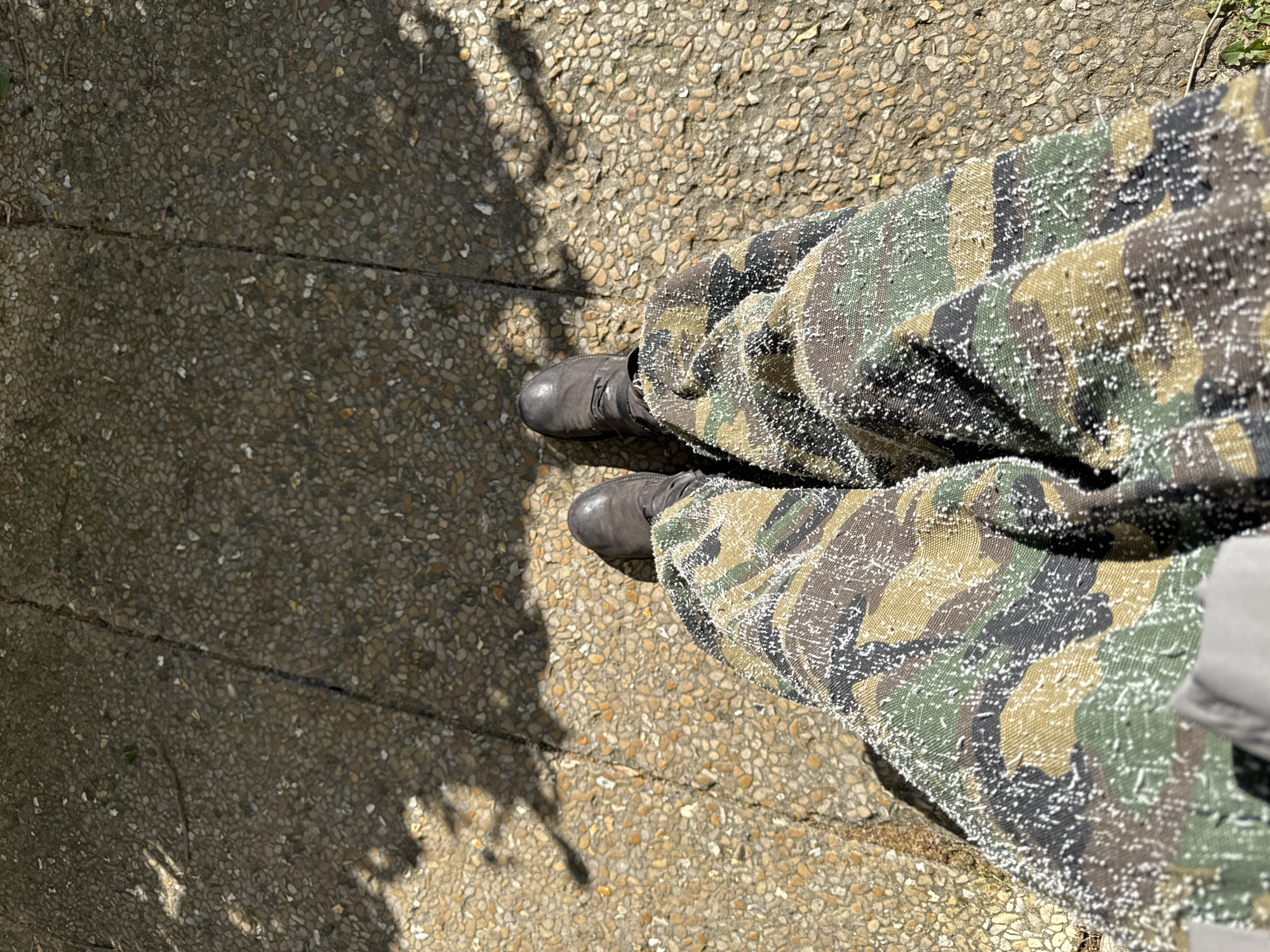 The image shows a top-down view of a person standing on a paved surface. The person is wearing camouflage pants covered with small, white particles. The pants are wide-legged and appear to be made of a textured fabric. The person's feet are clad in gray boots. A shadow is cast on the paved surface to the left of the person. The paved surface is composed of small, irregular stones. The location is Les Ulis, France.