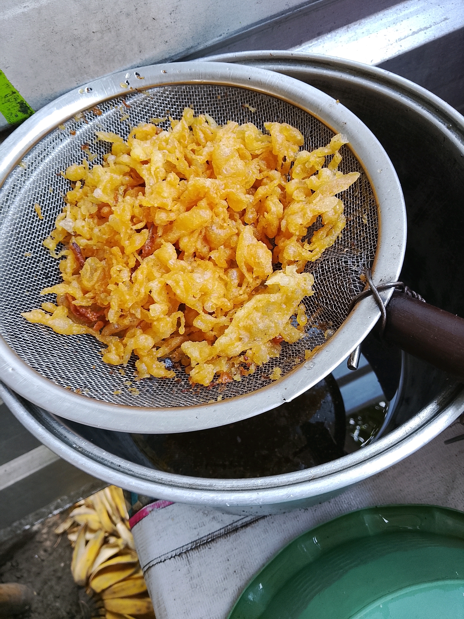 In Banaran, Indonesia, a close-up, overhead shot captures a batch of fried food, possibly banana fritters, resting in a metal sieve. The food is golden brown and appears crispy, with a batter coating. The sieve is positioned over a dark, oily liquid within a large metal pot. To the left, a glimpse of a grey, textured surface with a green stripe is visible, suggestive of a wall or building exterior. Below the pot, discarded banana peels are scattered on a dirt surface, next to a partially visible green plate. The lighting indicates an outdoor setting, possibly a street vendor's stall or a home kitchen area.