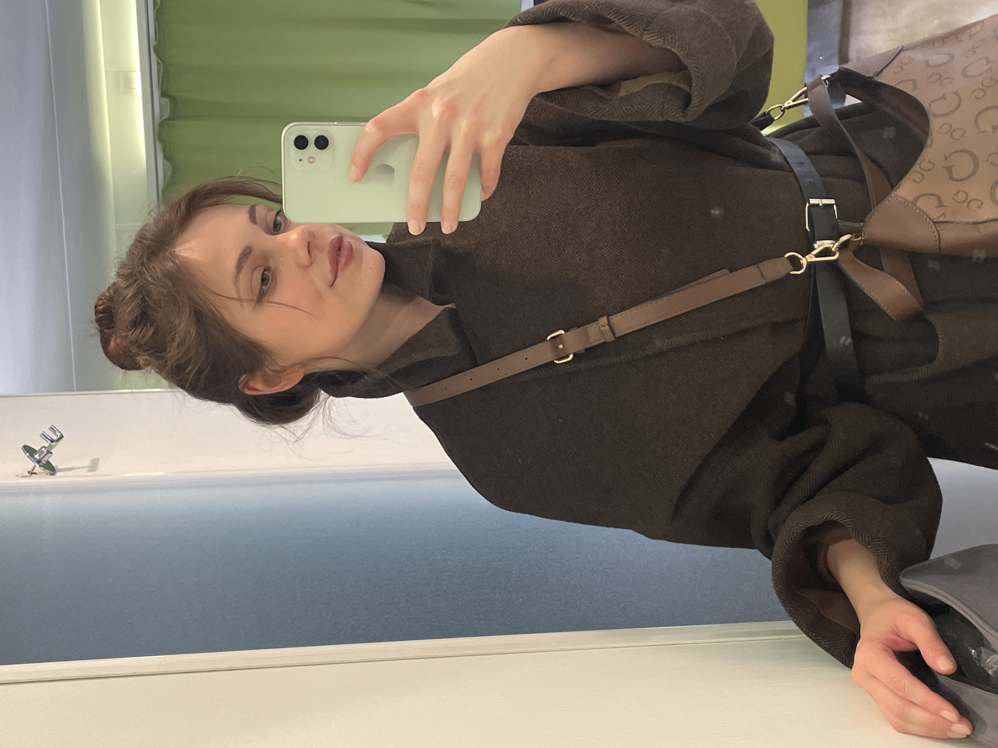 A young woman takes a selfie in a bathroom. She holds a light green iPhone in her right hand. Her hair is brown and gathered into a bun. She wears a brown tweed coat with a shoulder bag. The bathroom features a light blue countertop, a silver faucet, and a wall covered with green curtains. The location, Venice, Italy, is not visually discernible from the image.
