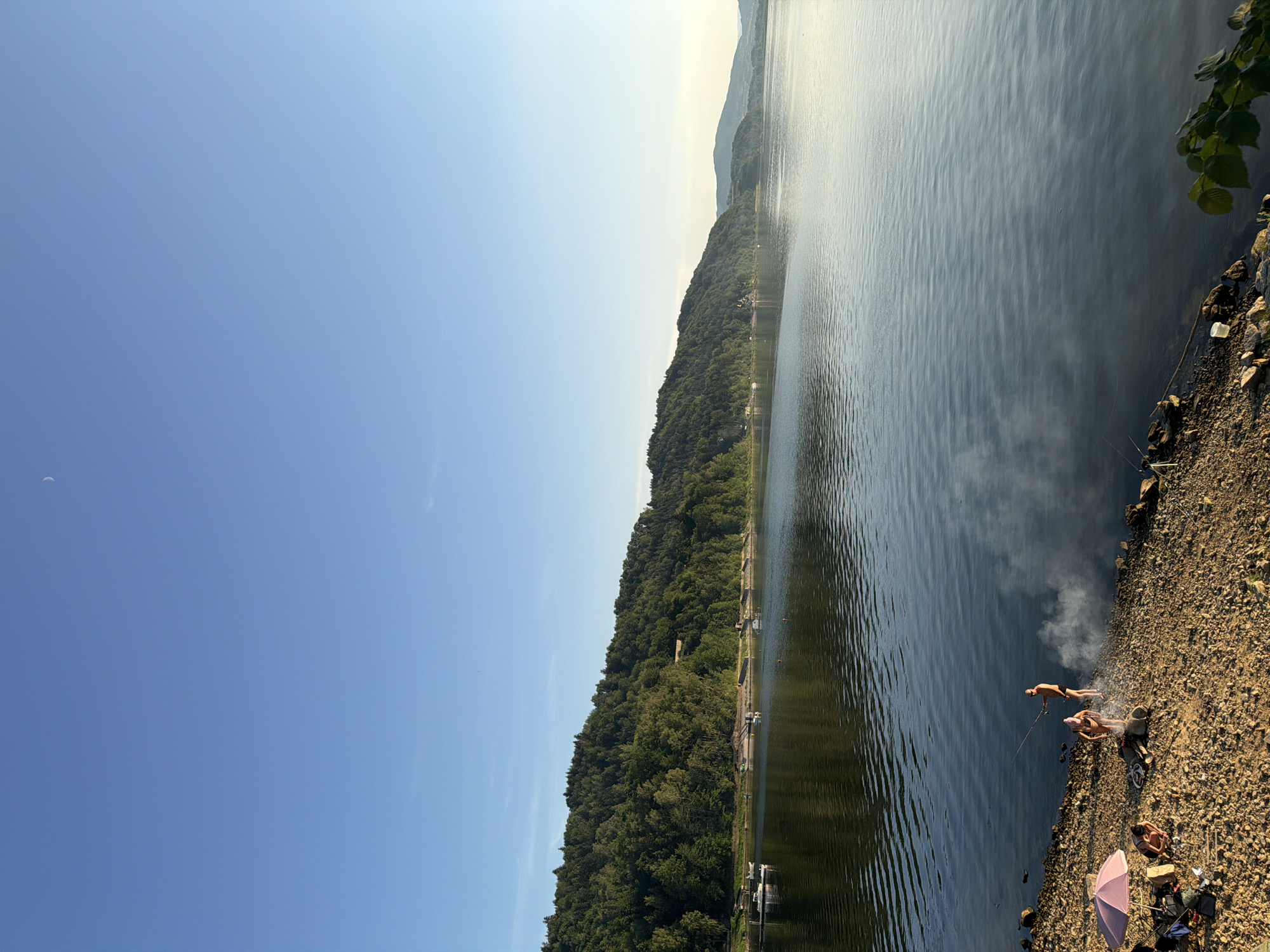 The image shows a serene landscape at the lake in Lipie, Poland. The clear blue sky is vast, with a faint crescent moon visible in the upper portion. A dense, verdant forest covers a rolling hill that slopes down to the water's edge. The calm lake's surface reflects the surrounding greenery and the sky.
In the foreground, a rocky shore is dotted with a few individuals engaged in recreational activities. Two people are standing near the water's edge, one appears to be fishing with a rod. A small campfire is burning, emitting a gentle plume of smoke. To the left, another person is seated on the shore, with a pink umbrella providing shade. Camping gear, including bags and a bicycle, are also visible on the shore. The overall scene depicts a tranquil day at the lake.