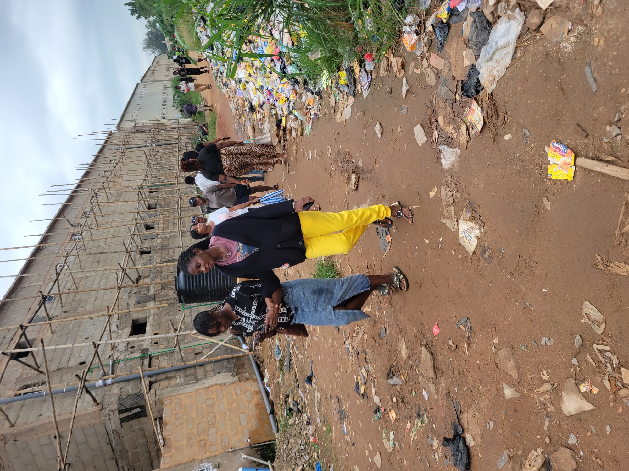 In Egbagu, Nigeria, a dirt path is lined with an abundance of scattered trash and debris, including plastic bags, wrappers, and what appear to be discarded food items. To the left, a long, unfinished building with a brick facade is under construction, supported by extensive bamboo scaffolding. A black water tank is visible against the building.
A group of people are walking along the path. In the foreground, two young women are walking side-by-side. The woman on the left is wearing a black and white patterned top and a long denim skirt, and is looking at her phone. The woman on the right is wearing a pink top and bright yellow pants, with a dark cardigan draped over her shoulders. Behind them, other individuals are also walking, some carrying bags. The overall scene depicts a community area with ongoing construction and significant litter accumulation.