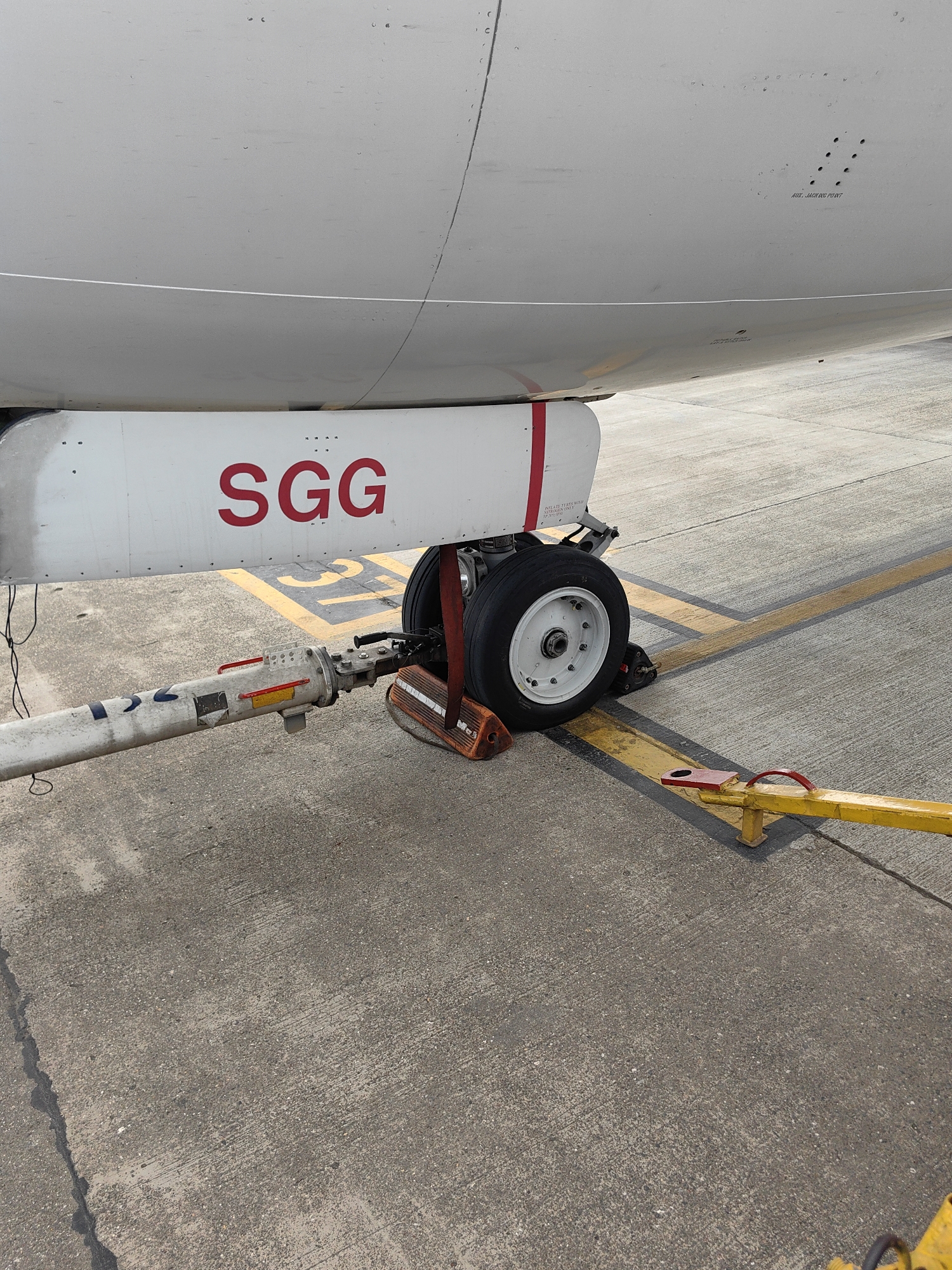 An airplane's nose gear is visible on a concrete tarmac, marked with yellow lines. The fuselage of the aircraft, painted white, is in the upper portion of the frame. The letters "SGG" are prominently displayed in red on a white panel attached to the aircraft's undercarriage. A refueling or service hose is connected to the nose gear, extending towards the left of the image. A yellow and red chocks are placed under the tire to prevent movement. In the background, a yellow tow bar is attached to the nose gear. The scene appears to be an airport tarmac, possibly in Mumbai, India.