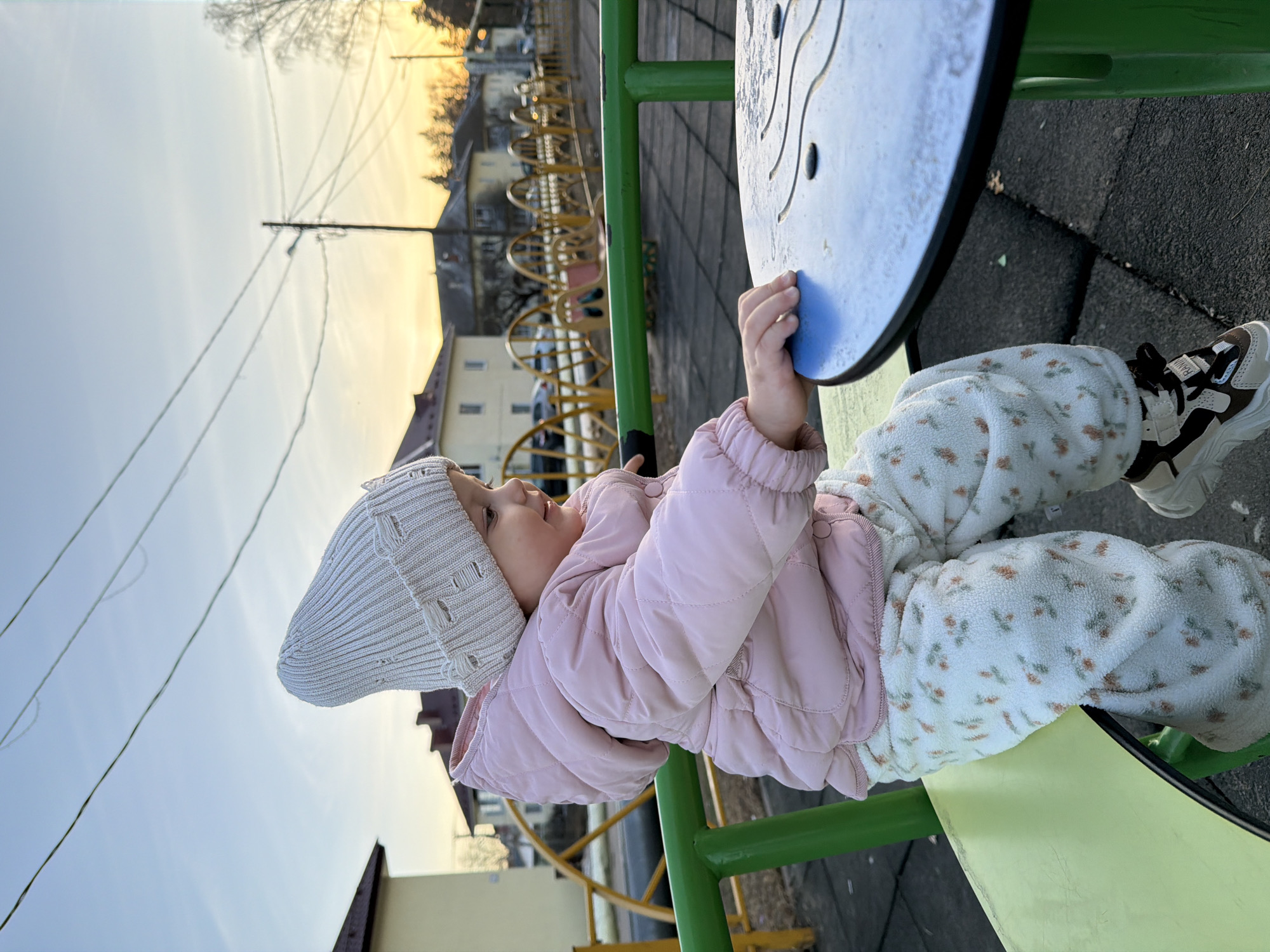 A young child, appearing to be under the age of five, is sitting on a playground structure. The child is wearing a light pink quilted jacket, floral-patterned pants, a beige knit hat, and brown and white sneakers. The playground structure features green metal bars and a blue circular surface with grooves. The background shows residential buildings, utility poles, and a yellow metal fence. The location is outdoors in Reshetnikovo, Russia.