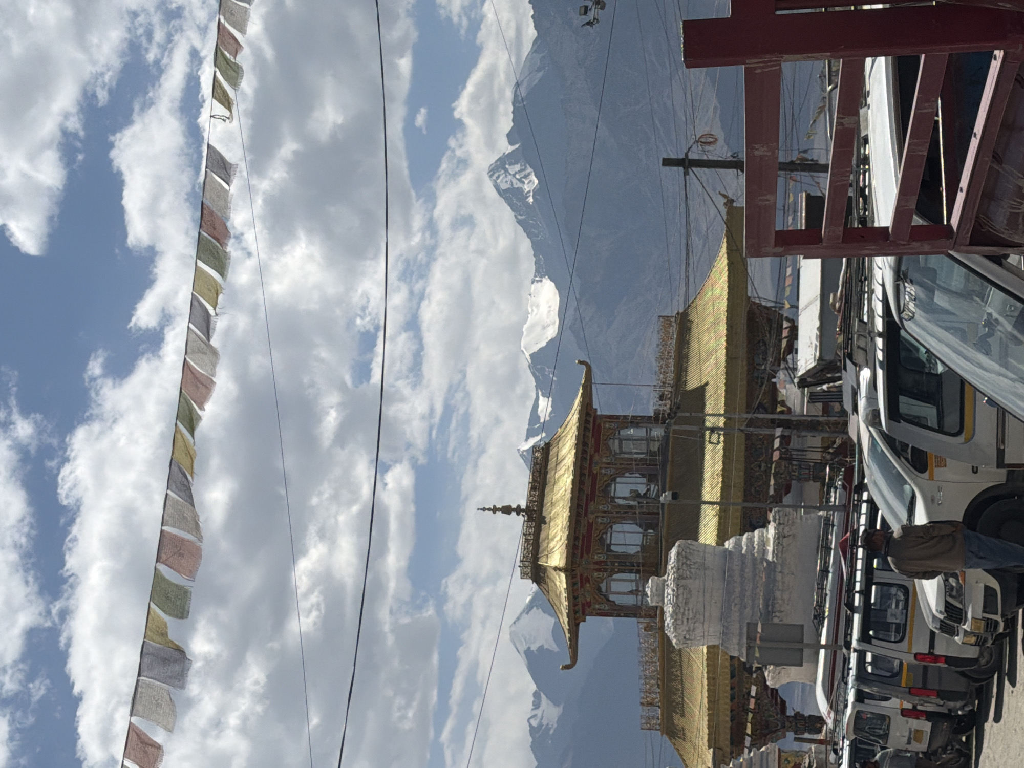 This is a photograph taken on a cloudy day in the city of Leh, India. In the foreground, a line of white vans is parked on a paved surface. A man wearing a brown jacket and blue jeans stands facing away from the camera, looking towards the vans. Behind the vans, a golden-roofed building with intricate red and gold decorations is visible. To the left of this building, a white stupa-like structure stands tall. In the background, majestic snow-capped mountains rise against a blue sky dotted with white clouds. Prayer flags, multicolored and rectangular, are strung across the sky, adding a touch of vibrant color to the scene. Electrical wires crisscross the frame, connecting various poles and structures. The overall impression is one of a vibrant cultural and natural landscape, typical of Leh.