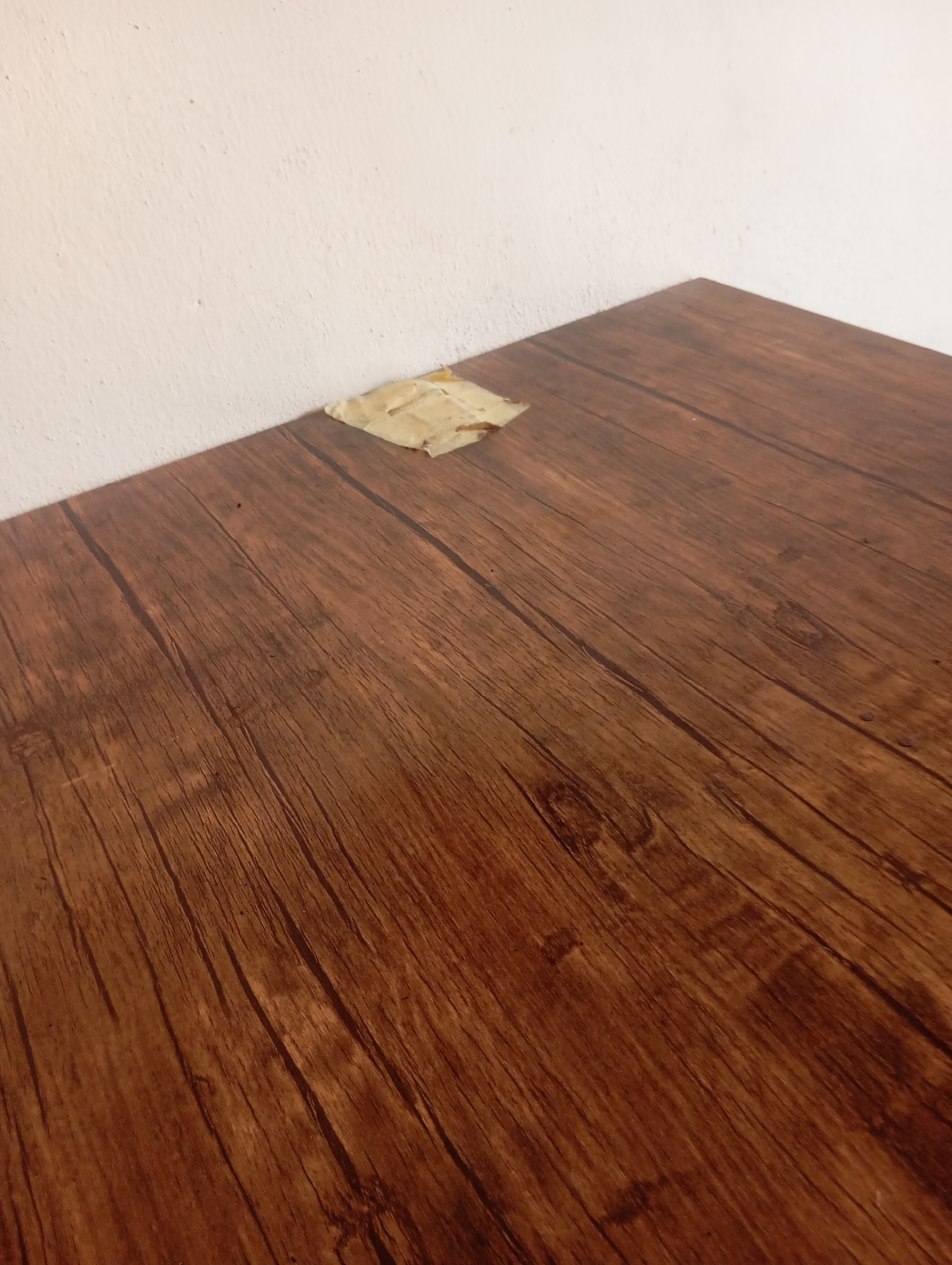A close-up, angled shot depicts a section of a brown, wood-grained laminate surface, possibly a table or countertop, against a textured, off-white wall. In the upper left quadrant of the laminate surface, a small, crumpled piece of beige tape is visible. The wood grain on the laminate runs diagonally from the bottom left to the top right of the frame. The lighting is dim and appears to come from the left, casting subtle shadows. There are no people or any other discernible objects in the scene. The location in the city of Jauro Yinu, Nigeria, cannot be determined from the visual information provided in the image.