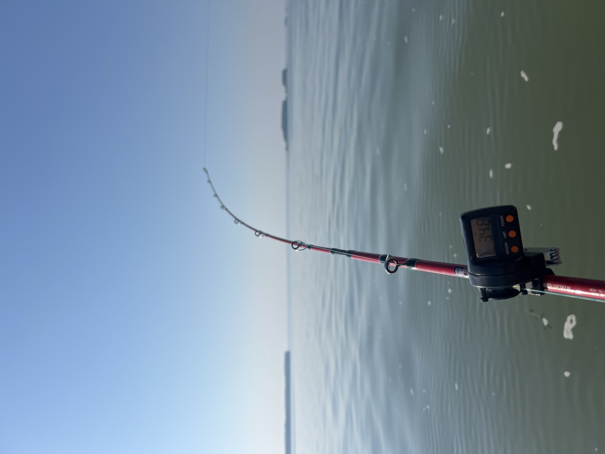 The image shows a fishing rod with a digital counter attached, angled upwards towards a clear blue sky and calm water. The counter displays the number "34.8". In the distance, there is a landmass, possibly an island or the shoreline, with a hazy horizon. The water is a muted green and reflects the sky, with small white foam patches visible on the surface. The fishing rod is red with black and silver accents, and it has several metal eyelets. The overall scene appears to be a serene outdoor setting, likely a body of water where fishing is taking place. There are no people visible in the image. The location is specified as Novopichugovo, Russia, suggesting this is a scene from that area.