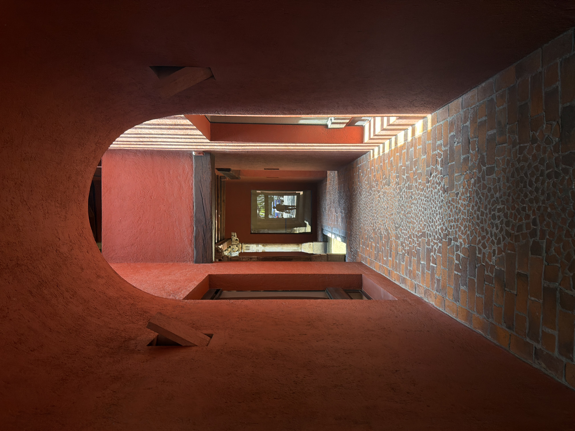 The image depicts an upward-angled view inside a building in Puebla, Mexico. The architectural style is characterized by earthy tones of red and orange, with textured surfaces. On the left, there is a curved wall with a circular opening. The right wall features a brick pattern. In the distance, there's a window. No people are visible in the image.
