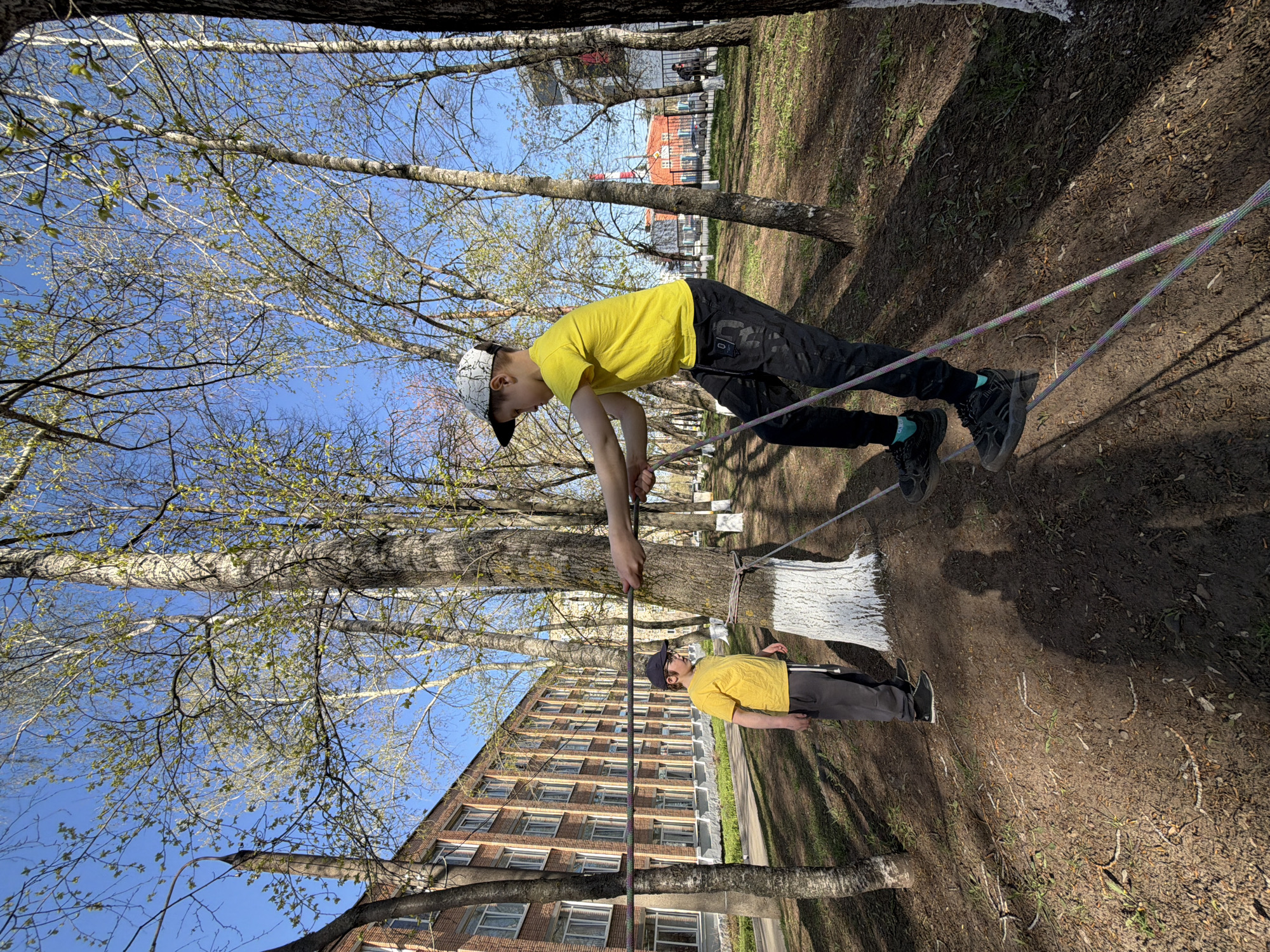 A photograph depicts a young male balancing on a slackline strung between trees in a park-like setting. The individual wears a yellow t-shirt, dark pants, a baseball cap, and dark footwear. He extends his arms for balance. The trees are bare, with a few nascent leaves. The slackline is multi-colored. In the background, there is a multi-story brick building. Based on visual landmarks and architecture, this scene is located in Izhevsk, Russia. An additional boy stands under the slackline wearing similar clothing. The ground is earth covered with fallen leaves and grass. The image is rotated to the side, creating a disorienting effect.
