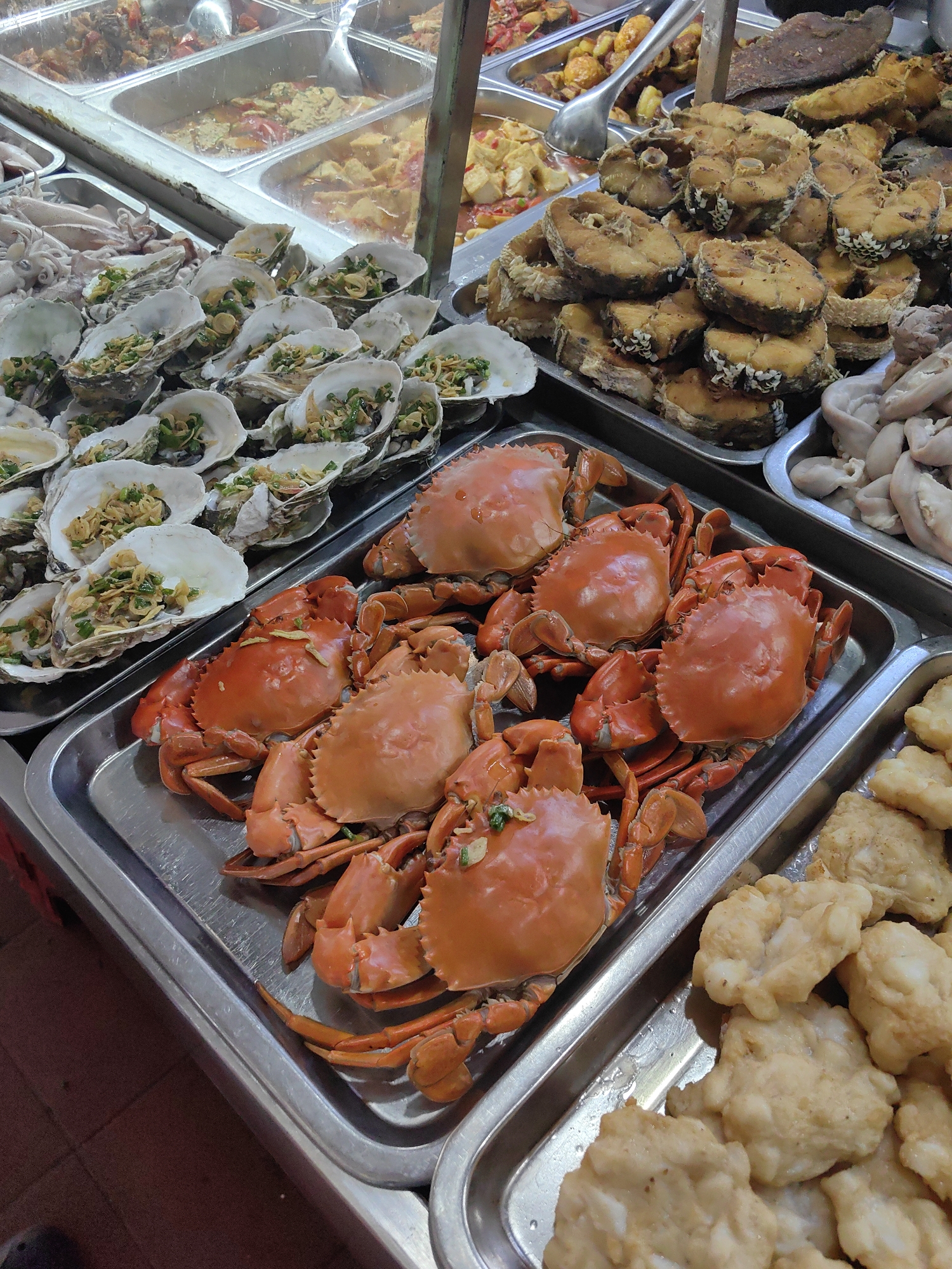 The image depicts a food stall in Hải Phòng, Vietnam, showcasing various seafood dishes. In the foreground, a metal tray holds several cooked crabs, their shells a vibrant orange-red. Adjacent to the crabs are trays of fried fish and oysters on the half shell, topped with herbs. Other trays contain fried foods and unidentified items. The scene is brightly lit, suggesting an open-air or well-lit indoor market environment.