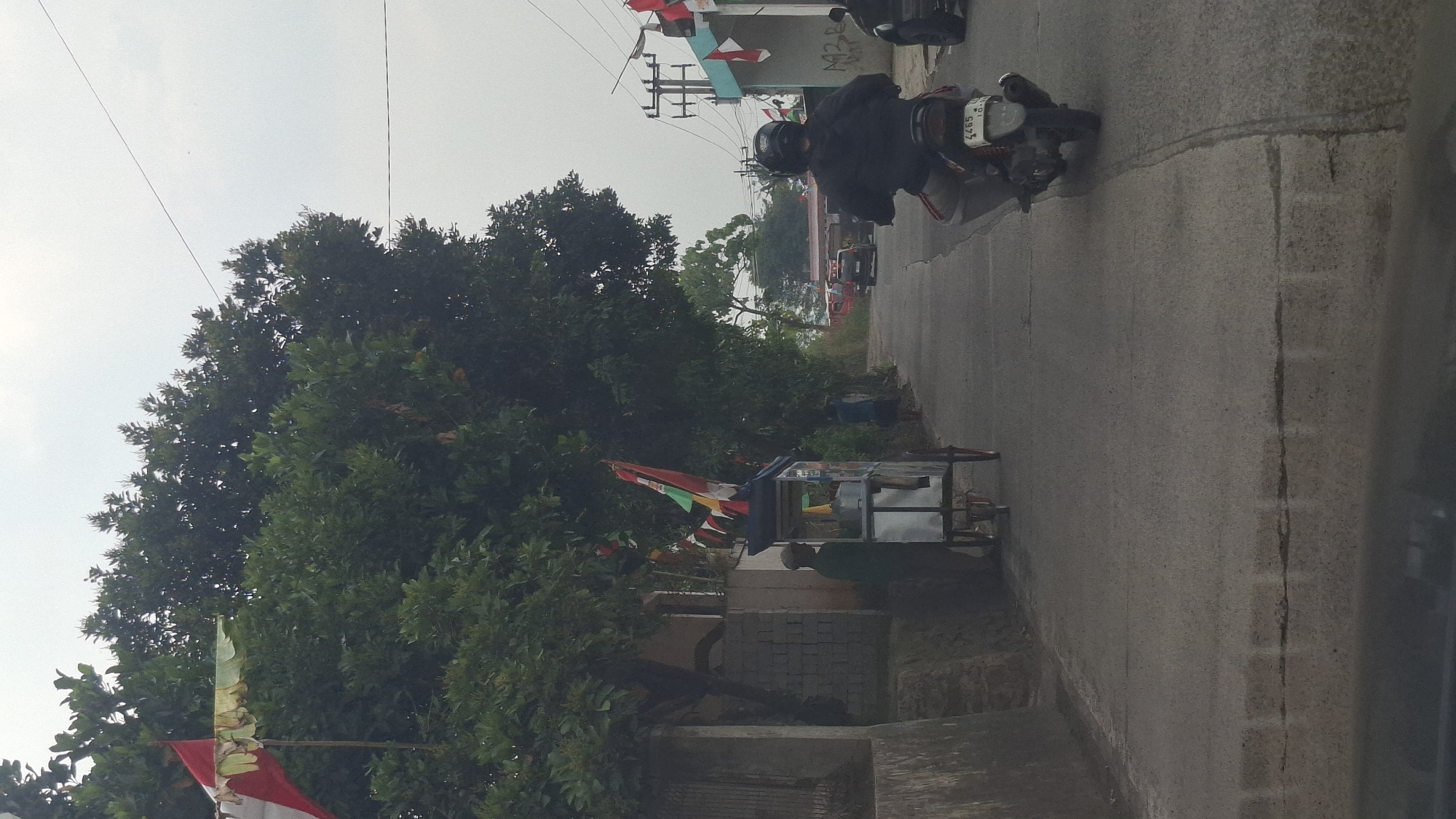 The image captures a street scene in Cibinong Hilir, Indonesia, under an overcast sky. On the left side of the road, a man stands beside a small, wheeled cart adorned with flags. The cart appears to be a mobile vendor. Behind the cart, lush green trees line the side of the road. The road itself is paved and appears to be a two-lane street. In the distance, several vehicles, including a red car and a black SUV, are visible. A person on a motorcycle, wearing a helmet and a dark jacket, is heading away from the viewer. Further down the road, a building with a blue and white facade is decorated with numerous flags. The overall impression is of a bustling street in a tropical environment.