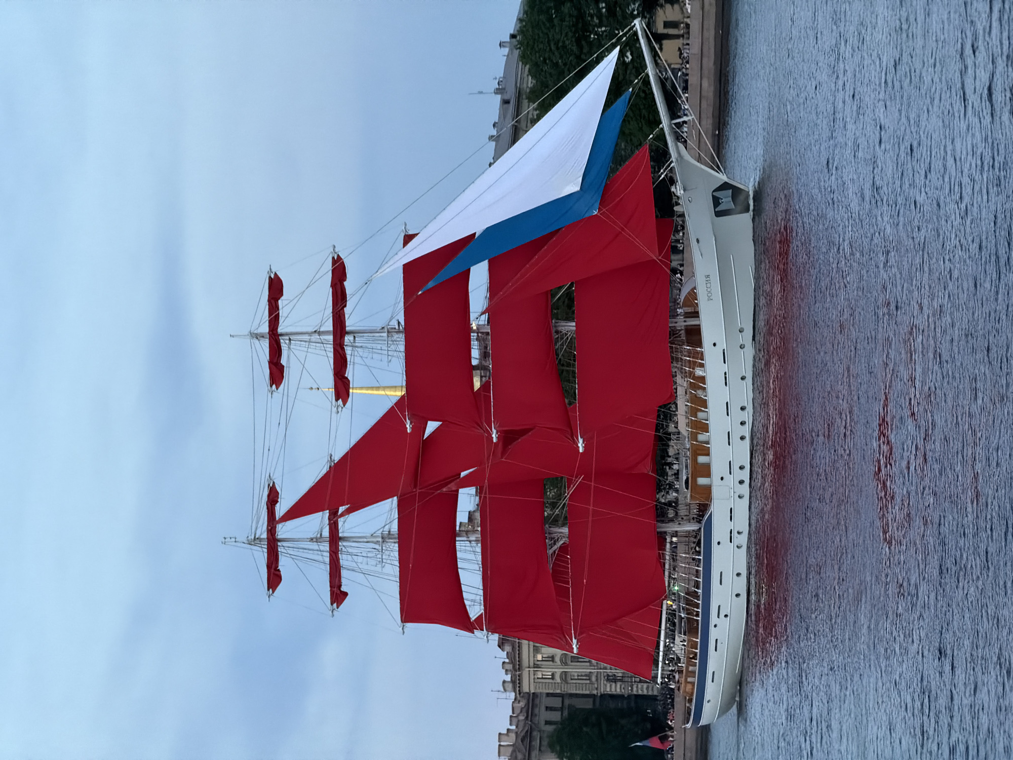 A large white sailing ship with vibrant red sails and a blue and white sail is sailing on a body of water in Saint Petersburg, Russia. The ship, named "РОССИЯ", is passing by a city waterfront lined with historic buildings. A crowd of people is visible on the shore. The sky is overcast and the water reflects the colors of the ship.