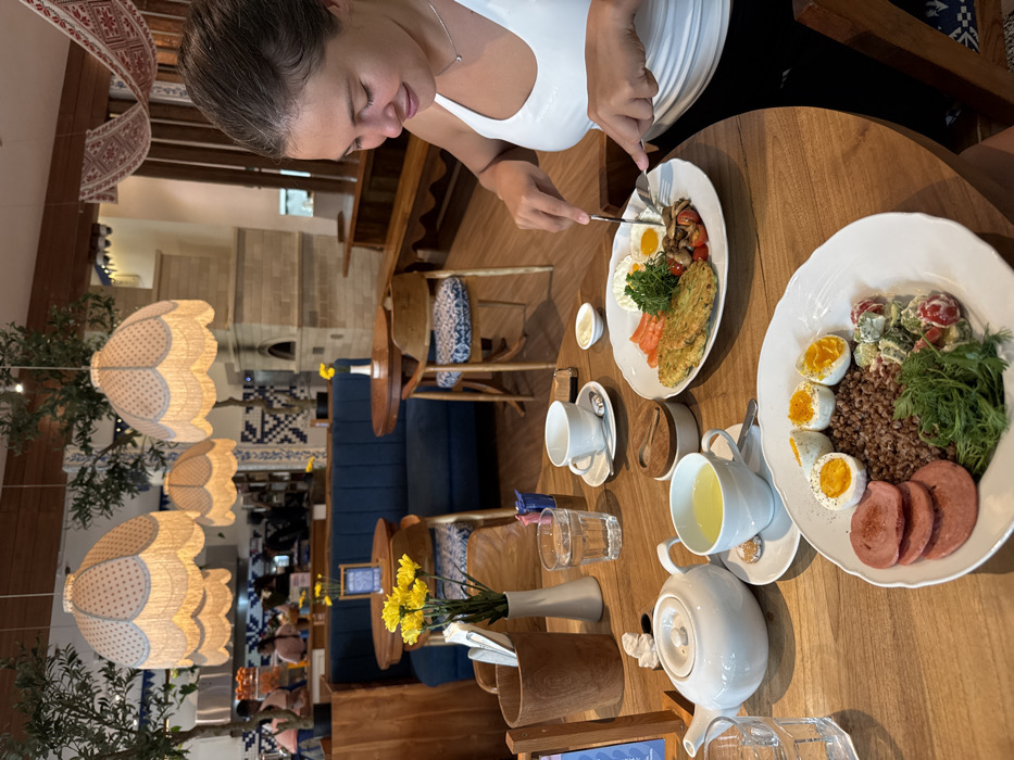 In a well-lit restaurant located in Kerobokan Kelod, Indonesia, a female individual is seated at a round wooden table, actively eating. She is light-skinned, with brown hair pulled back, wearing a white tank top. Her attention is directed downwards towards her plate as she uses a fork and knife to cut food.
On the table in front of her is a white plate containing two fried eggs, three large potato pancakes, a cluster of sautéed mushrooms, several cherry tomatoes, and a small portion of sliced smoked salmon or similar red fish, garnished with fresh parsley. Adjacent to this, closer to the viewer, is a second white plate featuring buckwheat groats, four halved soft-boiled eggs, three slices of cooked sausage, and a salad composed of diced cucumber and tomato with dill in a creamy dressing, topped with fresh dill.
Other items on the wooden table include a white ceramic teapot, two white ceramic teacups with saucers (one holding a yellow beverage), a clear glass of water, a small wooden container with a lid, a white ceramic creamer, a small white bud vase with yellow chrysanthemums, a wooden utensil holder, and a small wooden stand with a blue card.
The restaurant interior features a blue upholstered bench along the far wall, fronted by wooden tables and chairs with blue and white patterned cushions. Three large pendant lamps with scalloped, patterned fabric shades in white and light blue hang from the ceiling. Decorative trees in planters are positioned throughout the space. The walls exhibit white and blue patterned tiles. A prominent brick-faced oven or fireplace structure is visible on the right side of the background. Overhead and on the far wall, textile hangings with red and white patterns are draped. In the distant background, behind the blue bench, several individuals are partially visible at a service counter, some appearing to be staff.
