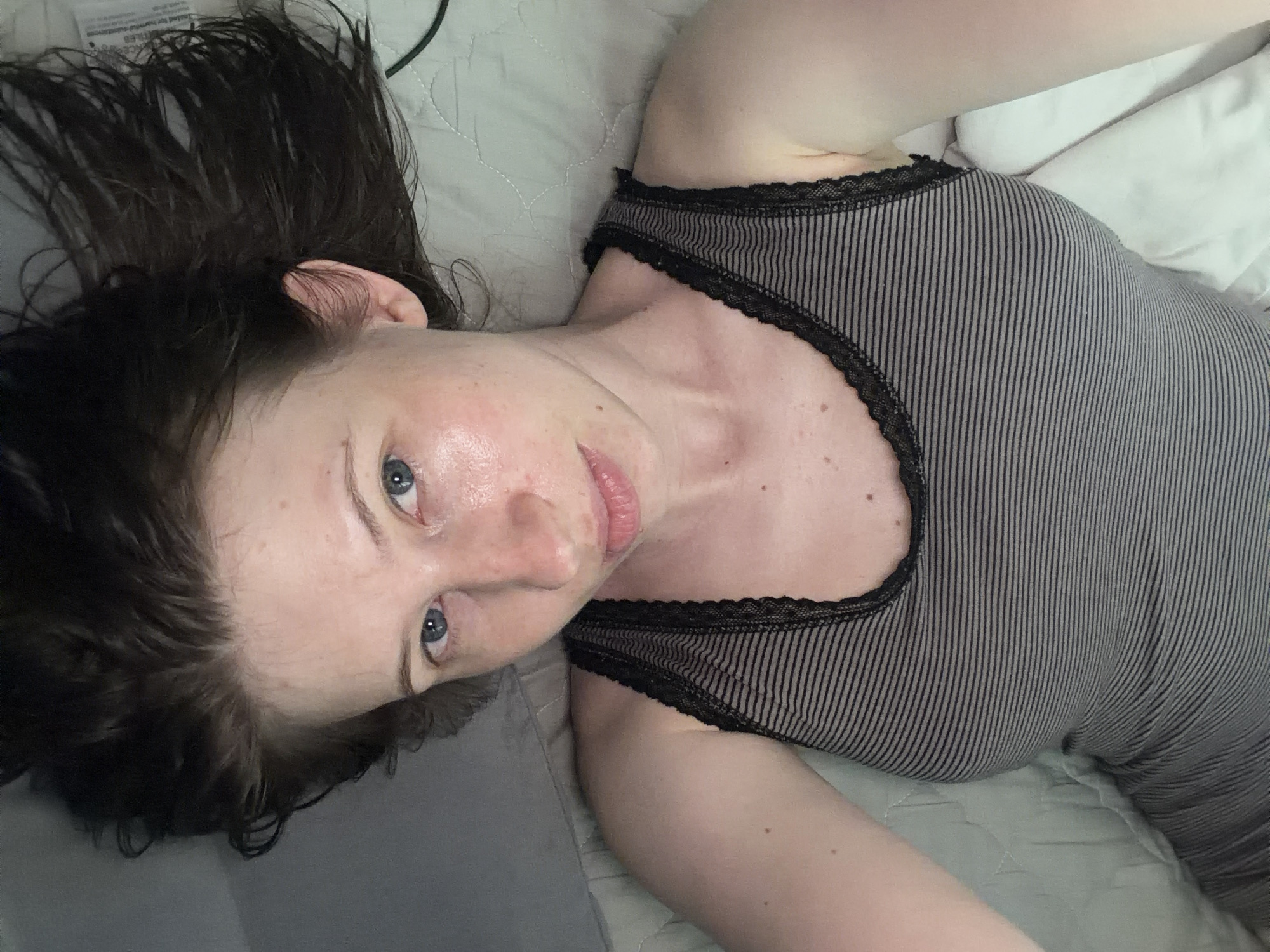 A person lies on their back on a bed, with their head facing the bottom of the frame and their feet towards the top. They are wearing a grey and black striped tank top with black lace trim. Their arms are raised above their head, bent at the elbows. The background consists of a textured grey surface, likely a bedspread, with a white pillow visible in the upper left corner. The lighting is soft, creating a slightly glossy sheen on the person's skin. The image is taken from a high angle, looking down at the person. There is no discernible information about the location in Dongducheon-si, South Korea, or any specific actions or interactions occurring.