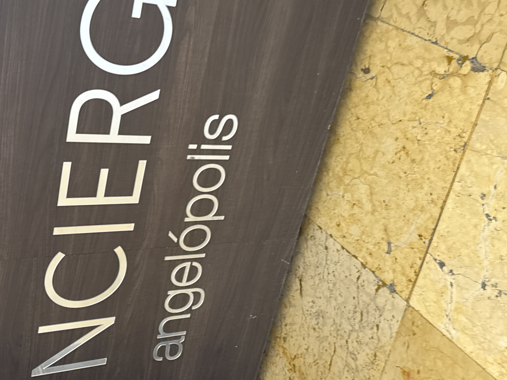 The image shows a portion of a dark gray sign with light gray lettering that reads "CONCIERGE" followed by "angelópolis". The sign is adjacent to a tiled floor composed of light yellow, roughly square tiles. The image suggests that the sign is located at a concierge desk in the Angelópolis area of Puebla, Mexico.