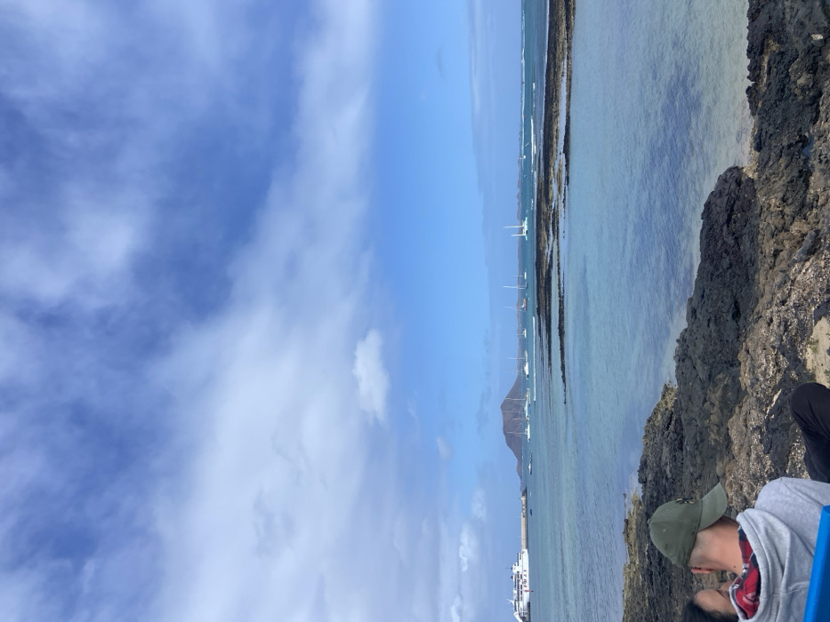 The image captures a view from a rocky shoreline in Corralejo, Fuerteventura, Spain (latitude: 28.737, longitude: -13.867). The scene features a vast expanse of blue water stretching towards the horizon, where several boats are anchored. The water is clear and turquoise, with a rocky seabed visible beneath the surface. In the distance, a mountainous landmass emerges from the sea. The sky is a vibrant blue, dotted with white, fluffy clouds. On the rocky shoreline, a lone figure sits with their back to the camera, wearing a green baseball cap and a blue hooded sweatshirt over a plaid shirt. The person appears to be gazing at the boats and the distant landmass. The scene portrays a peaceful and serene setting, offering a picturesque view of the coastal landscape.