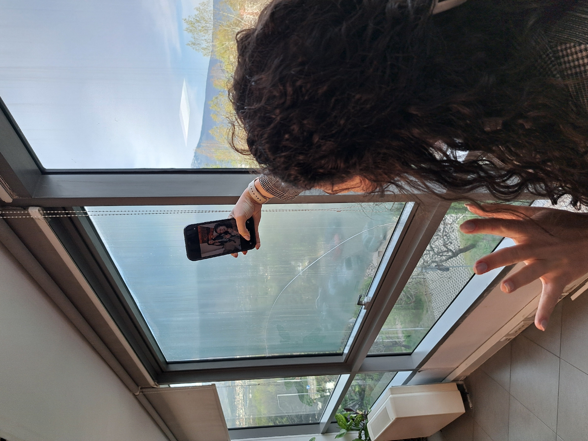 The image shows a person with curly dark hair holding a smartphone up to a cracked skylight. The phone screen displays an image of two people, possibly a photograph. The skylight is set within a gray frame. Visible through the skylight are trees and a distant landscape, suggesting an outdoor view. Another window is visible to the right with a different view of a tree and a building. There are some plants present near the second window. A hand is visible in the bottom right of the image. It is impossible to identify this location as Sofia, Bulgaria or any other location.