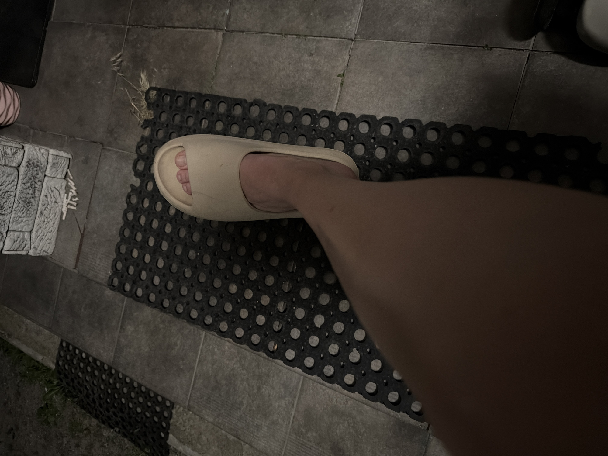 The image shows a person's bare foot, wearing a light-colored slide sandal, placed on a black rubber mat with circular perforations. The mat is situated on a tiled patio. In the background, there is grass, more tiling, and a portion of a light-colored brick structure. The lighting suggests it is either dusk or night. The scene does not provide any specific information to determine its location within Bila Tserkva, Ukraine.