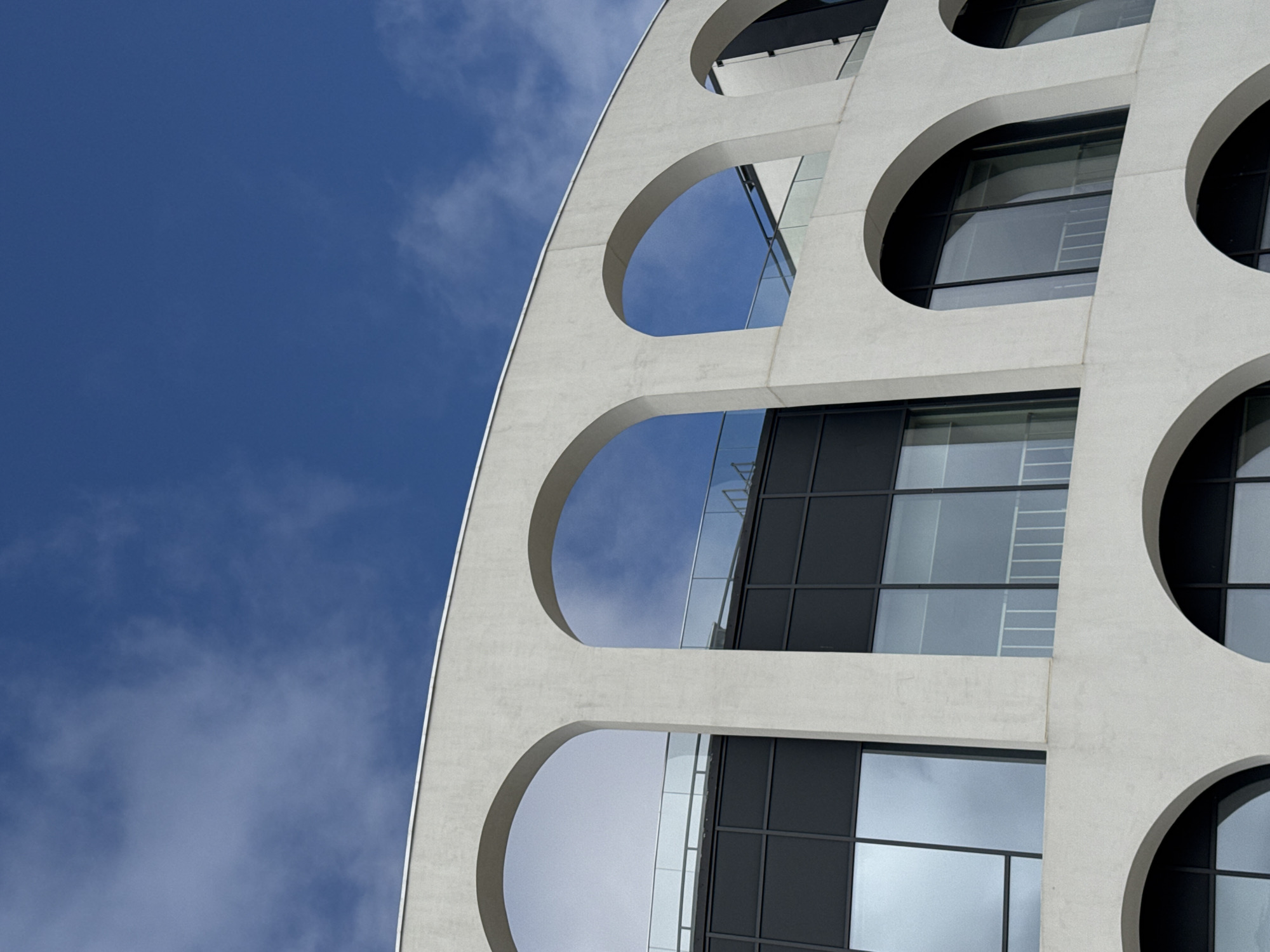 A low-angle shot reveals a modern architectural facade in Sochi, Russia, characterized by a series of semi-circular and circular openings. The facade is rendered in a pale, off-white material, likely concrete or a similar composite. The openings frame the building's inner structure, which consists of glass windows and dark-colored frames. A clear blue sky with wisps of white clouds forms the backdrop, visible through the openings and occupying a significant portion of the left side of the image. The composition focuses on the interplay between the building's design elements and the sky.