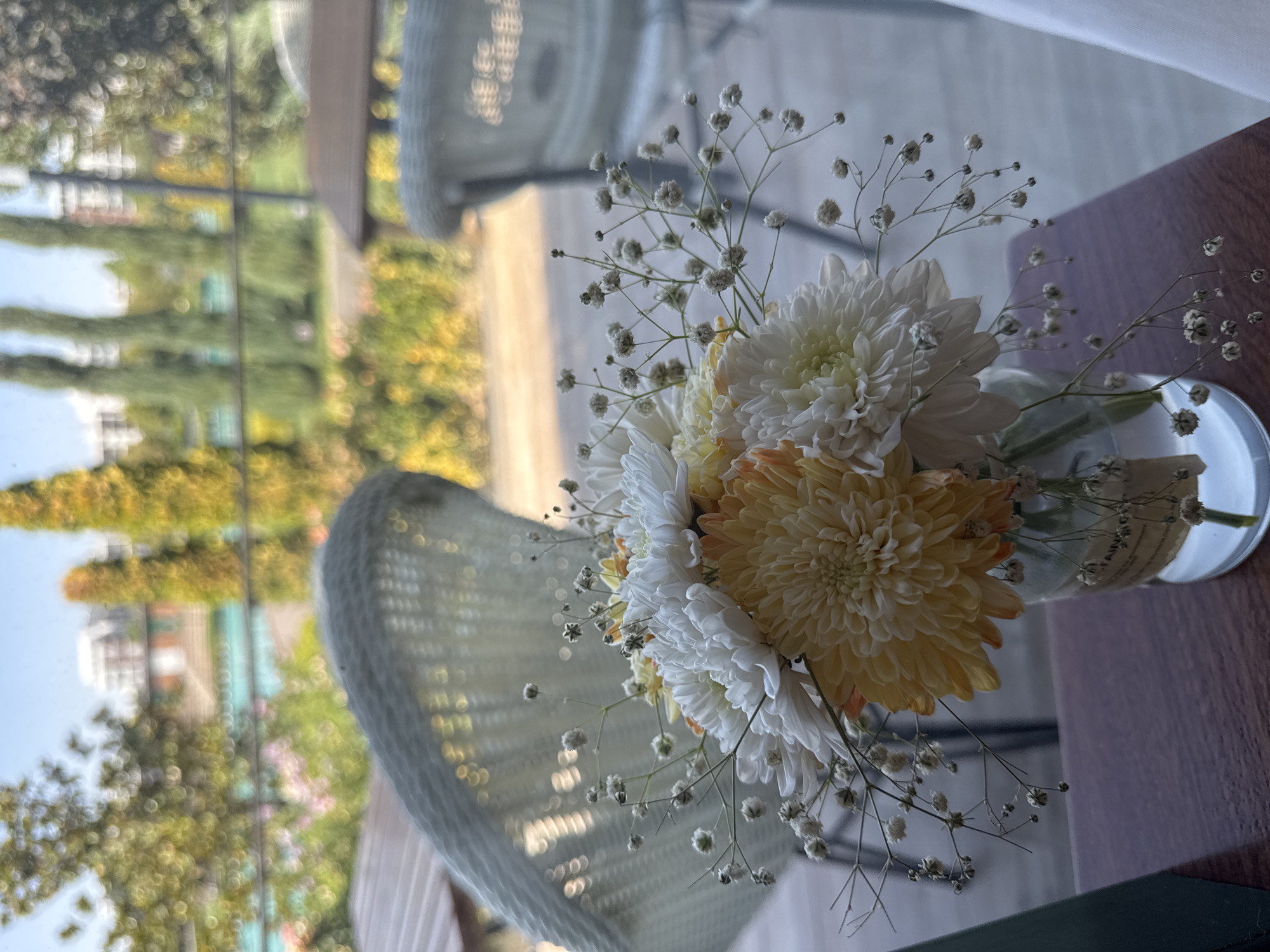 A close-up, eye-level shot focuses on a small bouquet of white and yellow chrysanthemums arranged in a clear glass vase. The flowers are delicate, with layers of petals, and interspersed with sprigs of baby's breath. The vase sits on a dark wooden surface, possibly a table. In the background, out of focus, is an outdoor seating area with wicker chairs and tables. Beyond that, the scene opens to a blurred view of a park-like setting with trees, foliage, and a body of water. The sunlight suggests it is daytime. The overall impression is one of a peaceful outdoor cafe or garden setting, likely in Tashkent, Uzbekistan, given the context of the query.