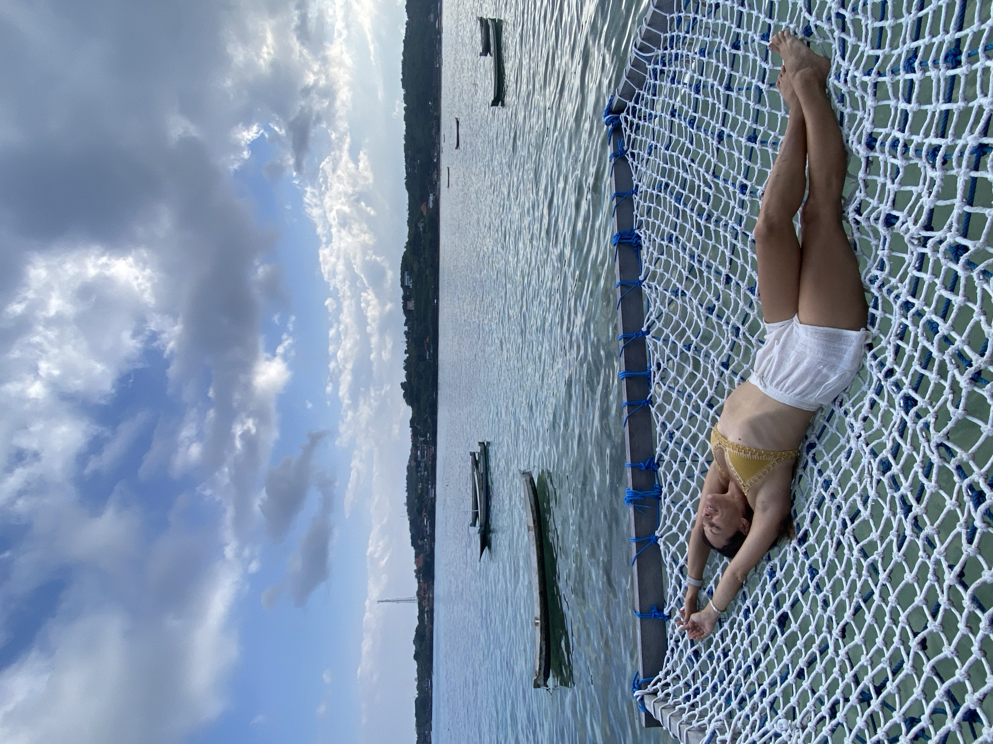 A woman is lying in a hammock suspended over the ocean. She is wearing a yellow bikini top and white shorts. Her arms are outstretched above her head and her eyes are closed. The hammock is made of white netting with blue ties. The ocean is calm and there are several small boats in the distance. In the background, there is a lush green hillside with some buildings on it. The sky is partly cloudy with scattered white and gray clouds. The scene is likely in Nusa Penida, Indonesia, a popular tourist destination known for its beautiful beaches and clear waters.