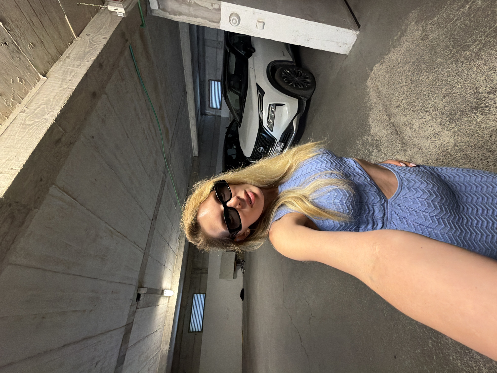A blonde woman, appearing to be in her 20s or 30s, is captured in a selfie within an underground parking garage located in Budva, Montenegro. She wears black rectangular sunglasses and a light blue knitted two-piece outfit featuring a chevron or zigzag pattern with a cutout on her left waist. Her long blonde hair is styled with a side part, falling over her shoulders. Her right arm is extended, holding the camera. The background consists of a utilitarian concrete parking structure. The ceiling displays raw concrete with visible wood grain patterns from formwork, intersected by concrete beams fitted with industrial fluorescent lighting. A green electrical conduit runs along one of the ceiling beams. On the left wall, a rectangular louvered ventilation grate is positioned high, above a low concrete barrier or ledge. To the right of the woman, a white Nissan SUV, identified by the Montenegrin license plate "07 BV 158", is parked, showcasing black multi-spoke wheels. A portion of a dark-colored vehicle is visible behind the Nissan. The concrete floor exhibits a textured and worn appearance.