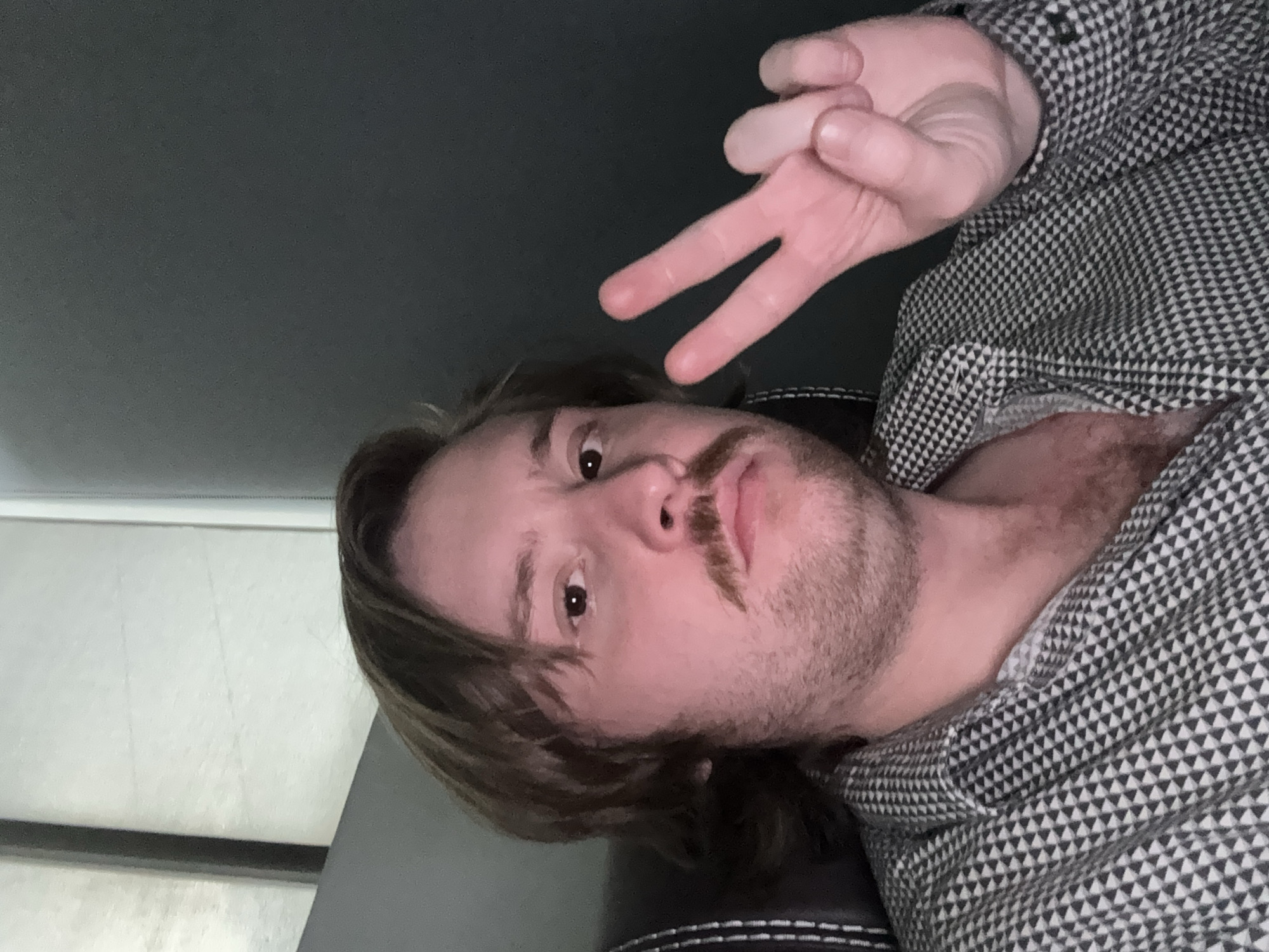 A man with brown hair and a mustache is lying down, facing the camera, and making a peace sign with his right hand. He is wearing a patterned black and white shirt. The background consists of a dark gray wall above him and what appears to be a light-colored surface below. The location is not discernible from the provided image.