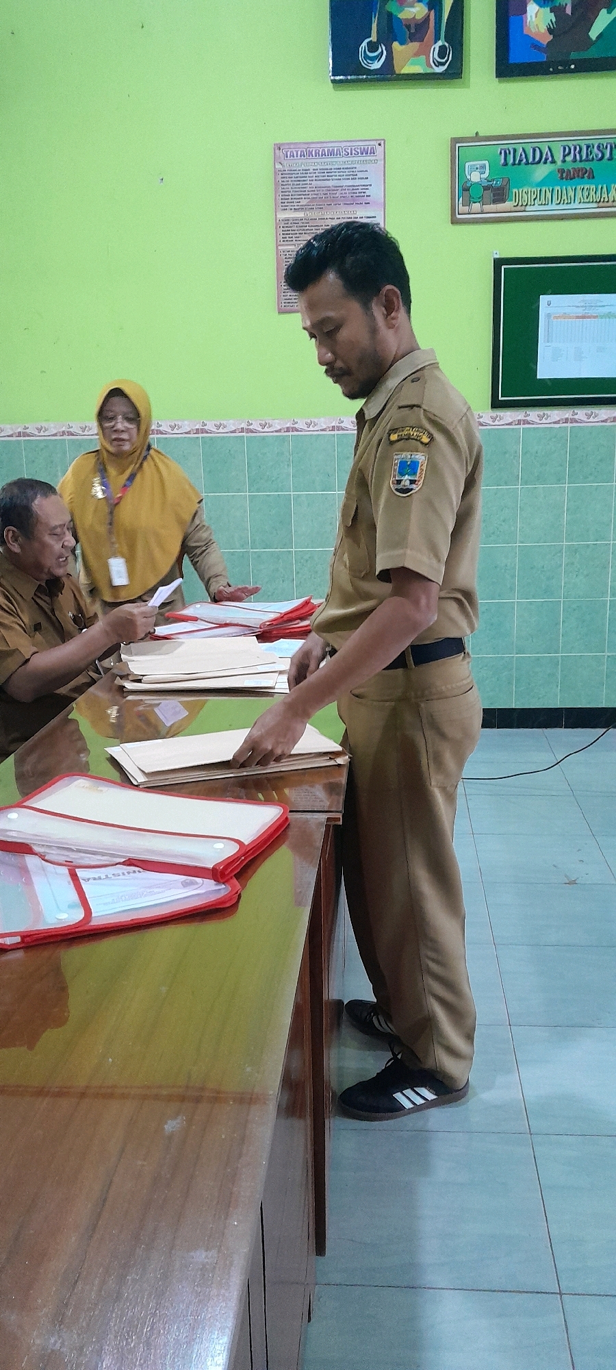 The image captures a scene inside a room, likely an office or classroom, in Tahunan, Indonesia. A man in a tan uniform, with a badge on his left sleeve, stands looking down at documents on a wooden table. He is holding a stack of folders. To his left, another person in a similar uniform is seated, also looking at documents and holding one paper. Behind them, a woman wearing a yellow hijab and a tan uniform stands observing. The room has light green walls and tiled flooring. Posters are visible on the wall behind the individuals, one titled "TATA KRAMA SISWA" and another with the text "TIADA PRESTASI TANPA DISIPLIN DAN KERJA KERA." A whiteboard or bulletin board with a document is also present on the far wall. The overall impression is of an administrative or educational setting where paperwork is being handled.