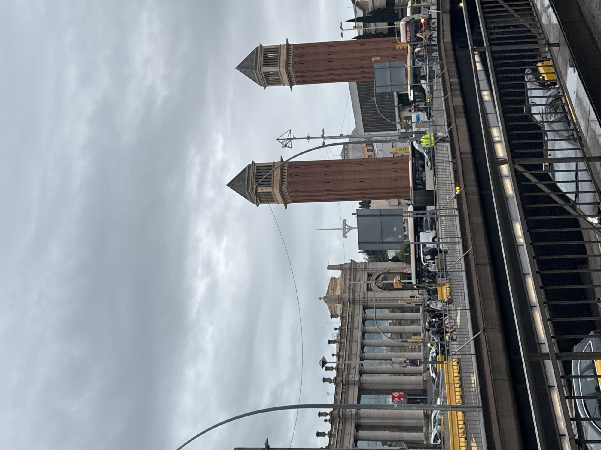 This photograph captures the city of Barcelona, Spain, under a cloudy sky. Prominent landmarks are visible: two brick towers with pyramid-shaped roofs stand out against the gray sky. In the foreground, there's a metallic structure, possibly an elevated pedestrian walkway or part of a transit system, with people moving along it. A classical building featuring numerous columns can be seen next to the towers. Cars and buses drive on the streets below.