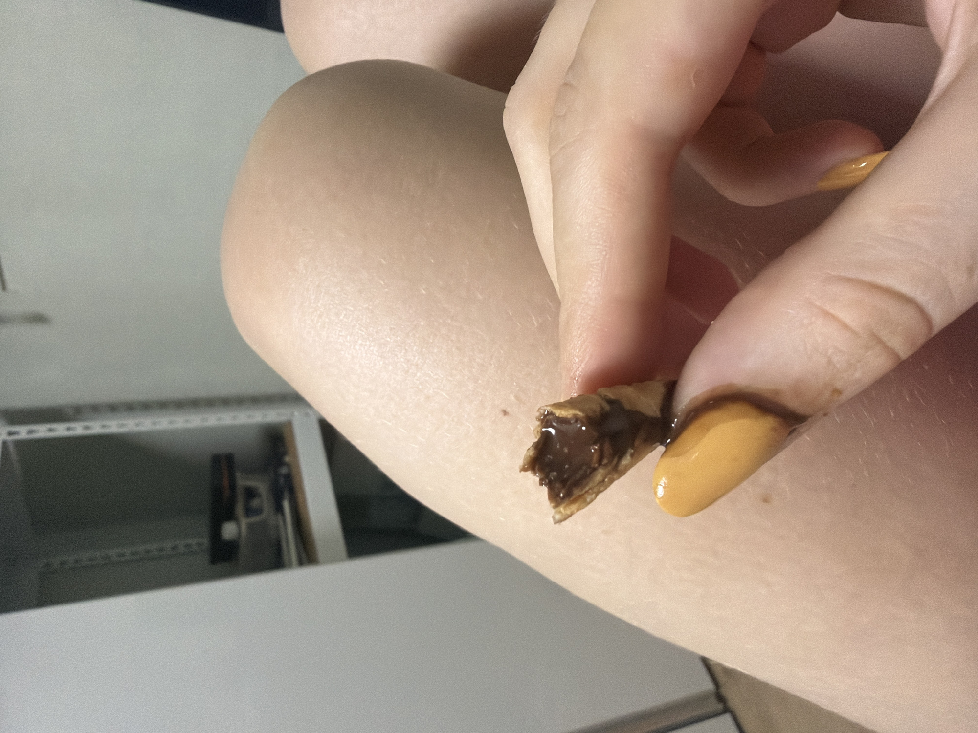 A person's hand is holding a partially eaten chocolate-filled wafer cone over their lap, which is covered by skin. The wafer is broken, revealing the chocolate filling. The person's fingernails are painted a bright orange color, and some chocolate has smudged onto their index finger. In the background, a white shelf unit with various items is visible. The scene appears to be indoors.